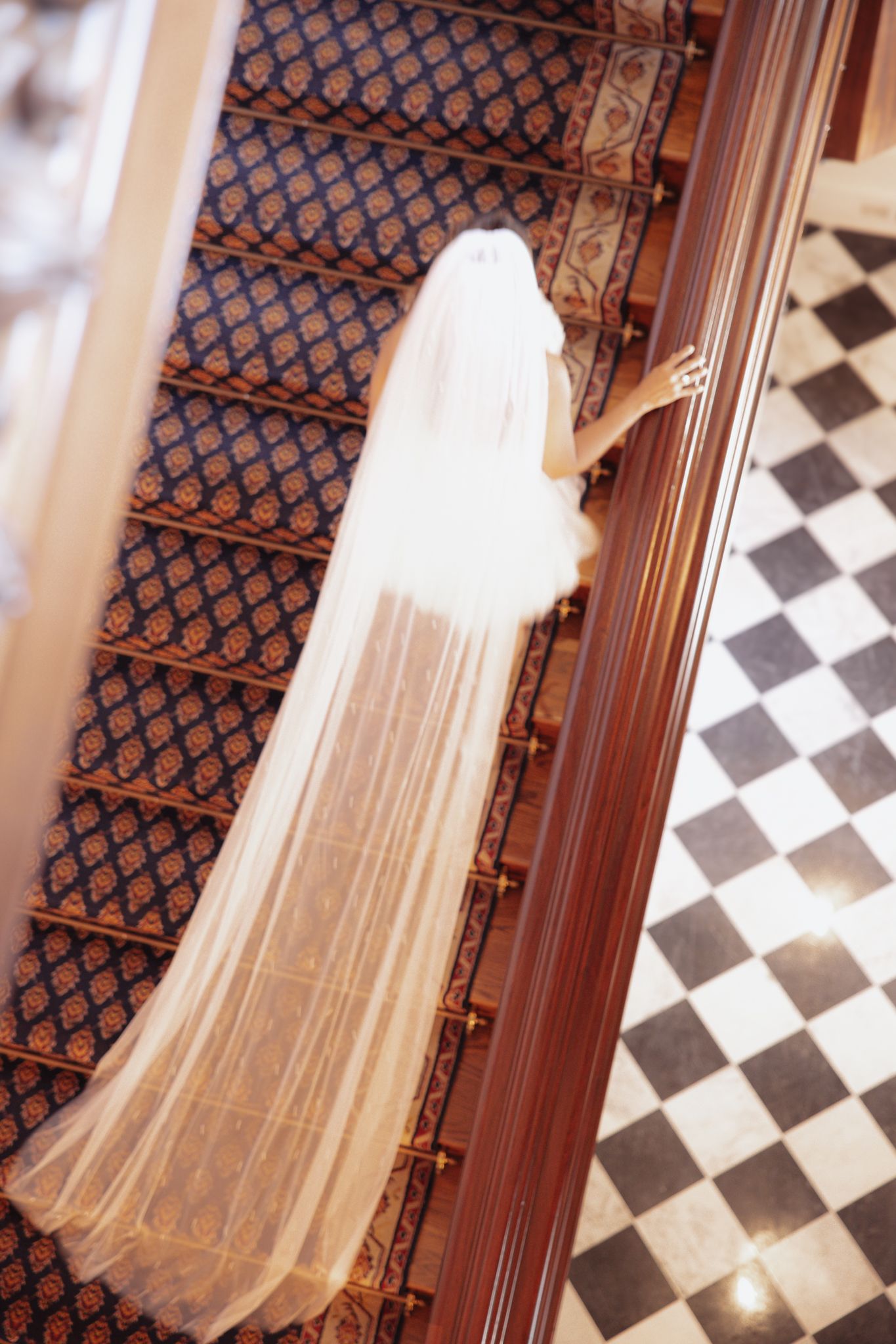 Flowing white bridal veil cascades down ornate staircase with geometric patterned carpet and checkered floor.