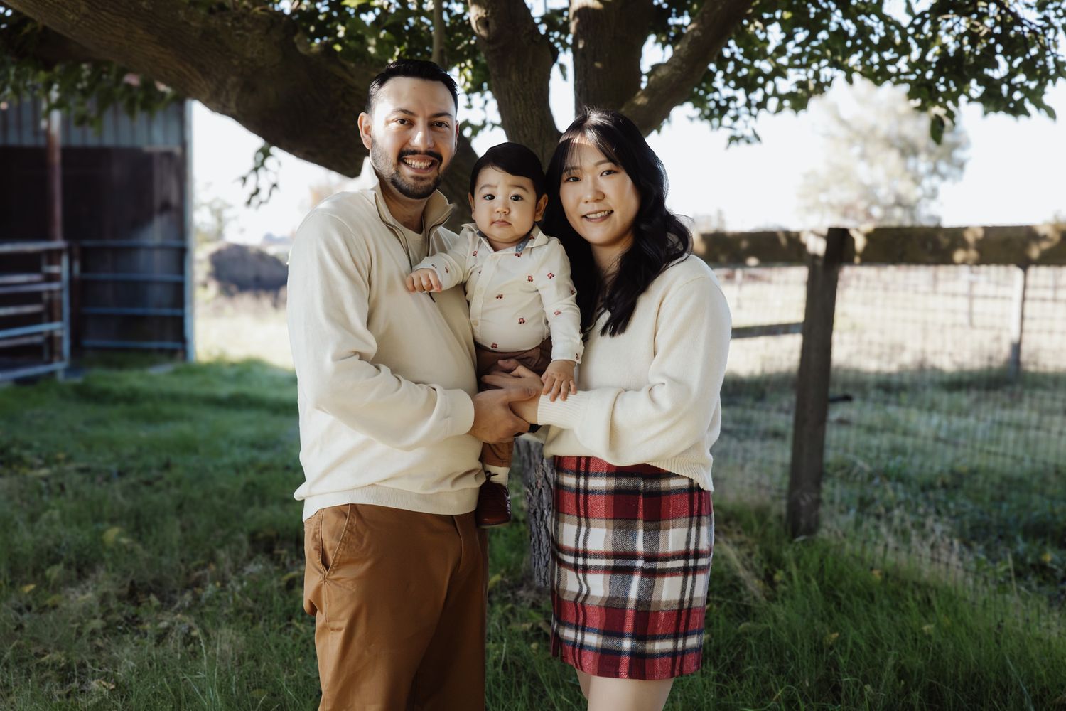 A family in coordinating cream and plaid outfits poses together in a grassy outdoor setting during golden hour.