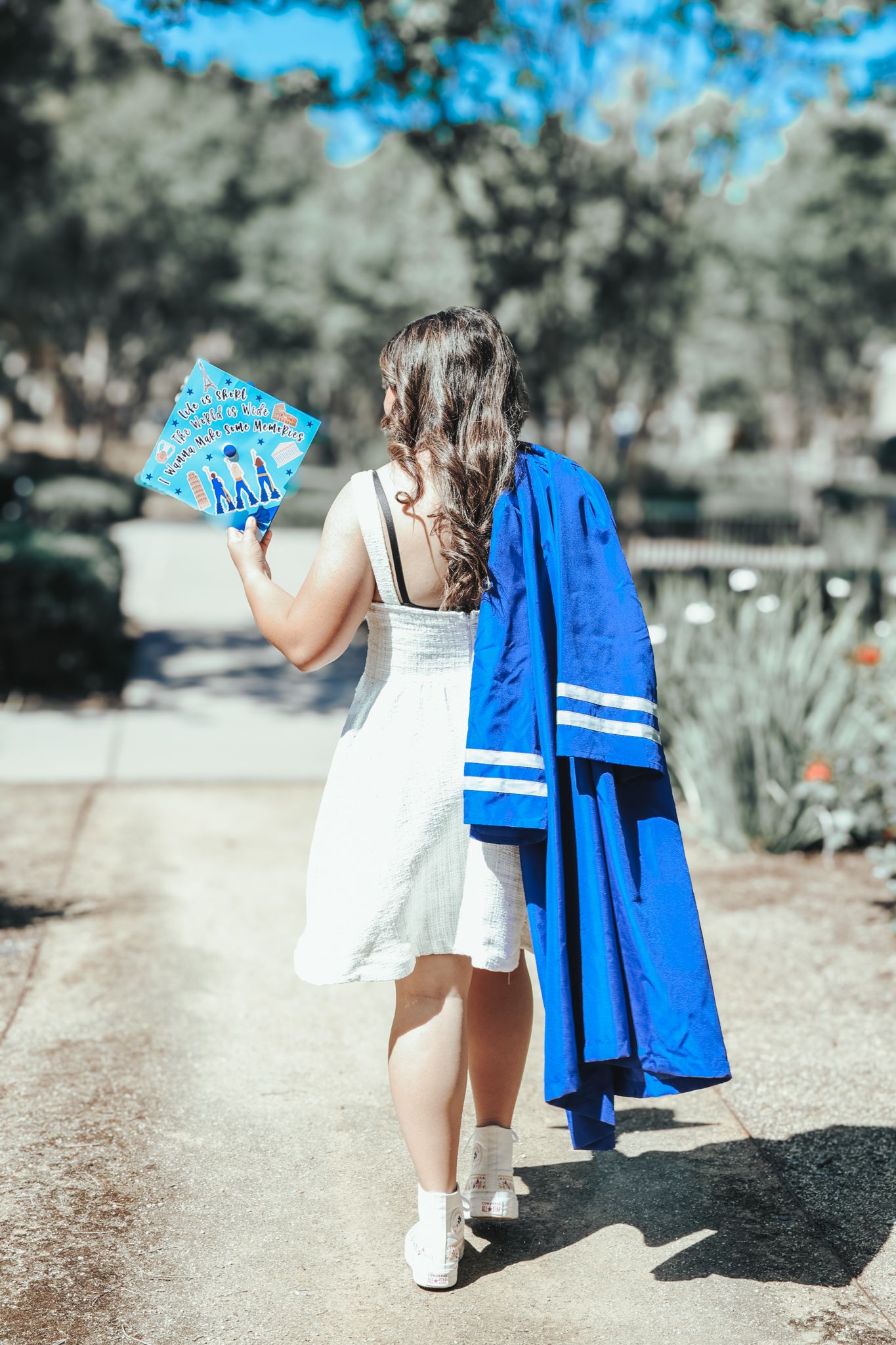 Two figures walking on path carrying blue graduation gowns on sunny day.