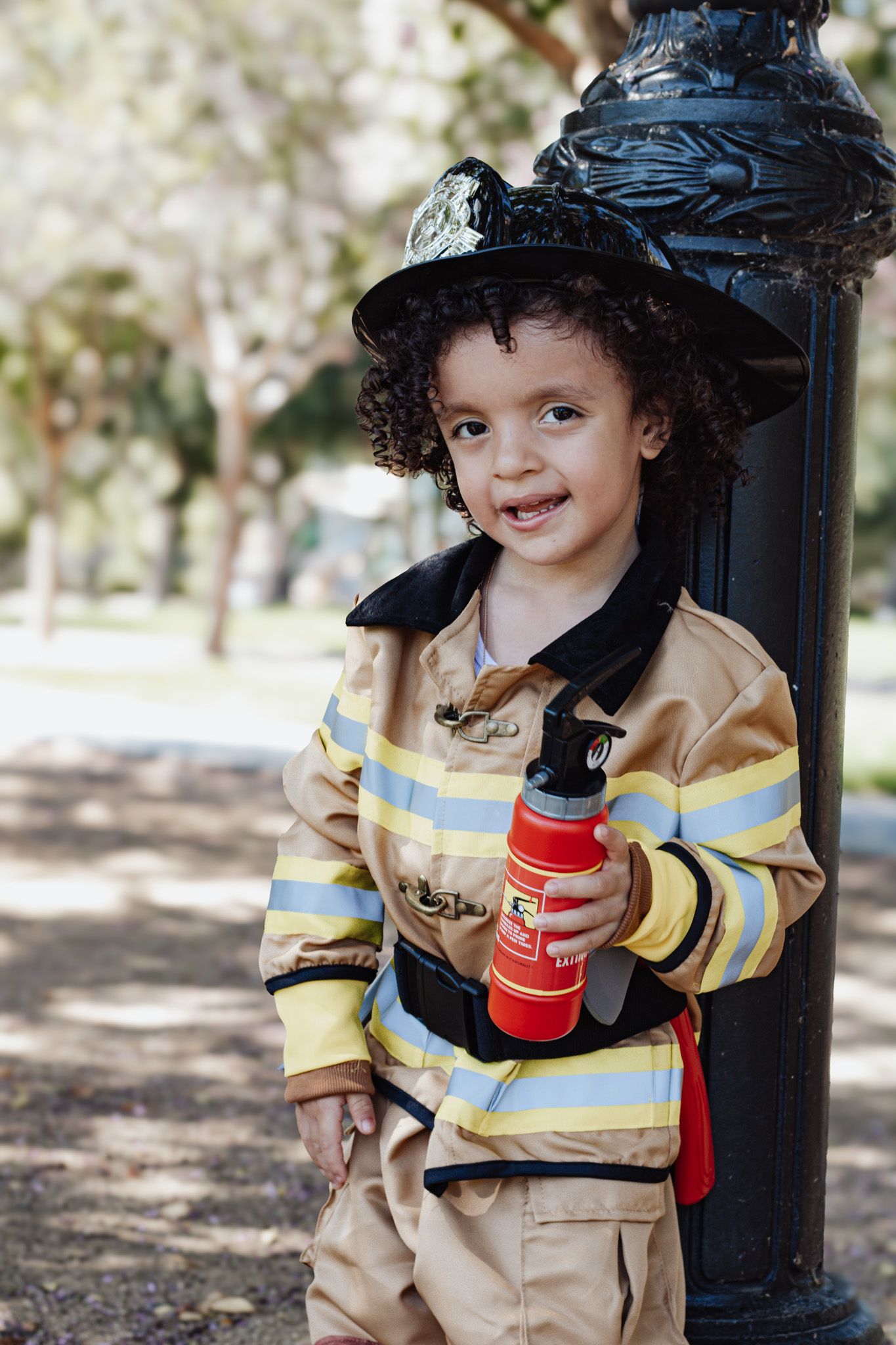 Smiling child in firefighter costume holding toy extinguisher, leaning against lamppost in park.