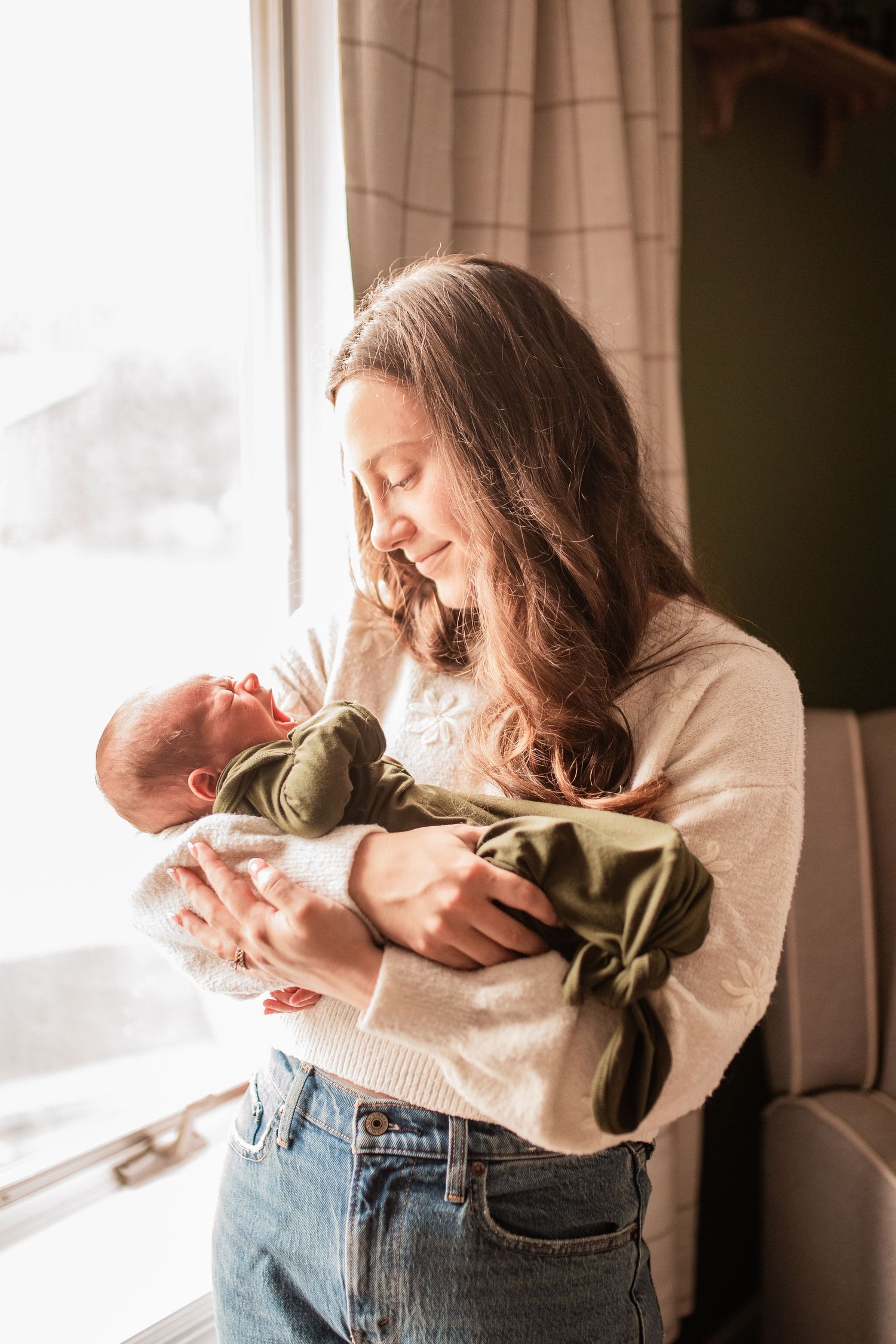 Natural light lifestyle photo of a mother holding her newborn baby by a window, capturing a tender motherhood moment. Schuylkill County