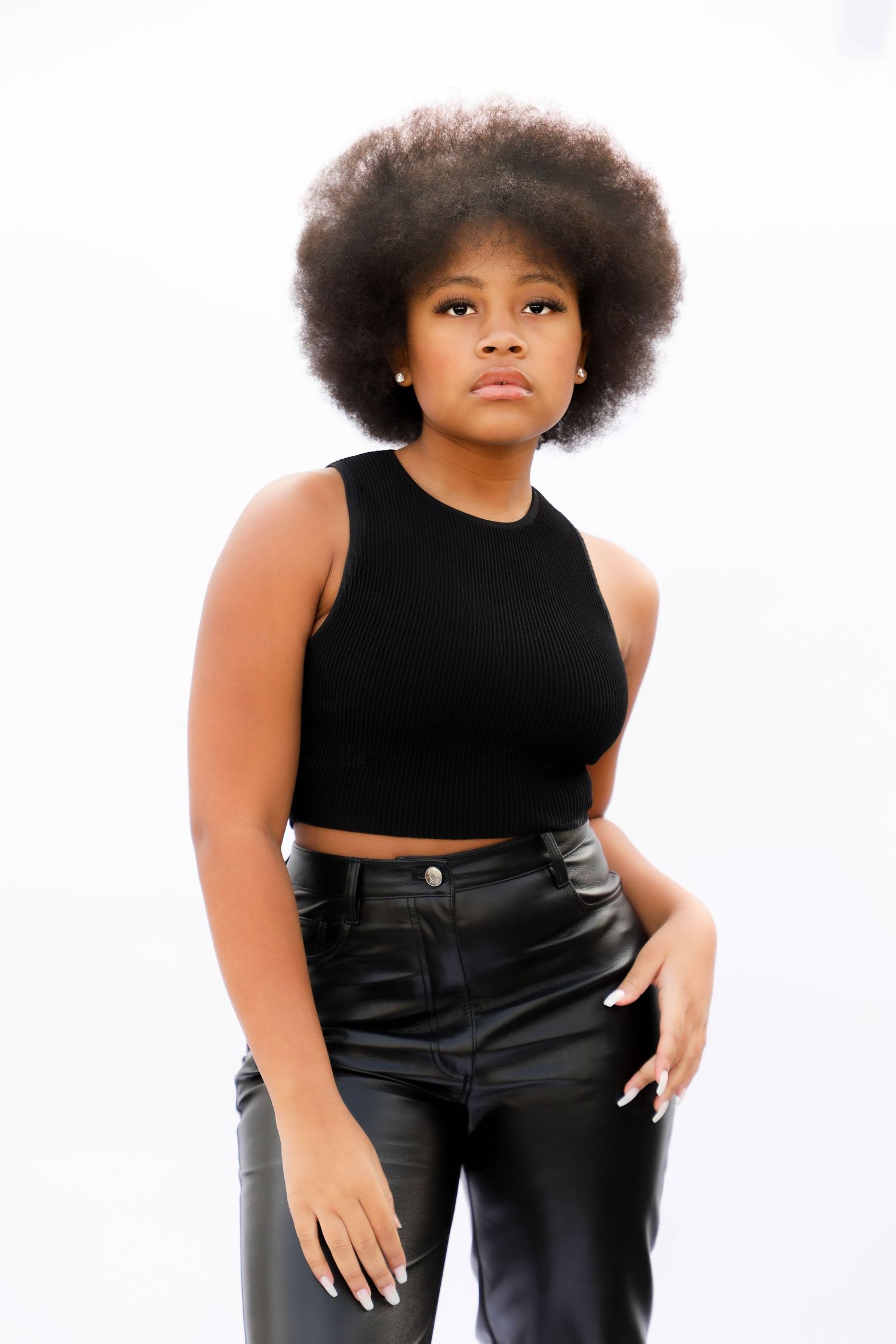Young Black woman with afro hair wearing black crop top and leather pants posing against white background.
