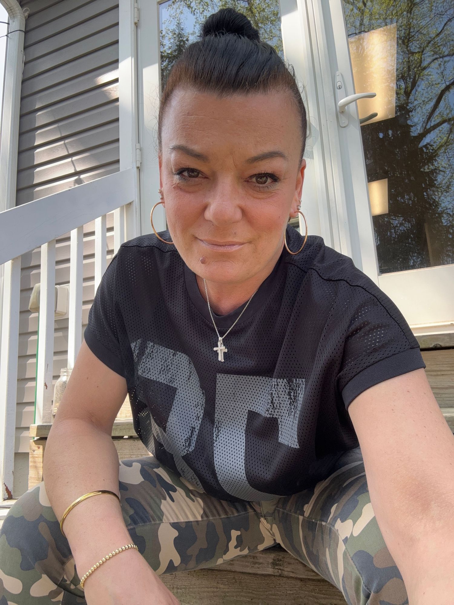 Woman in black graphic tee, camo pants, cross necklace, and hoop earrings sitting on wooden steps outdoors.
