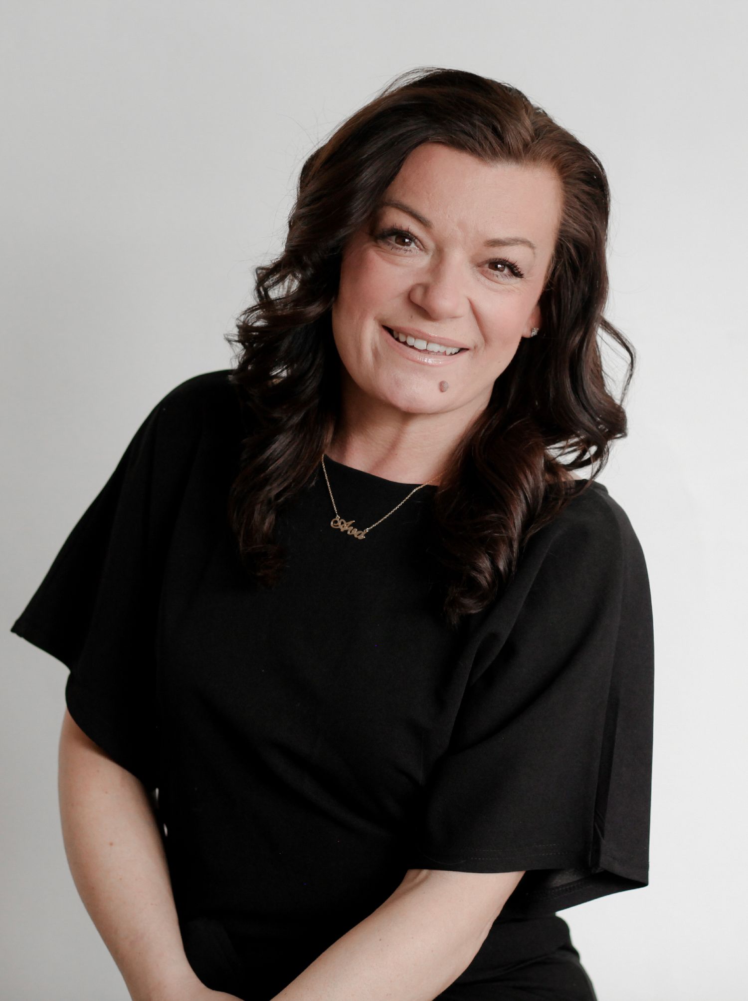 Smiling brunette woman in black blouse with gold necklace posing against light gray studio background.