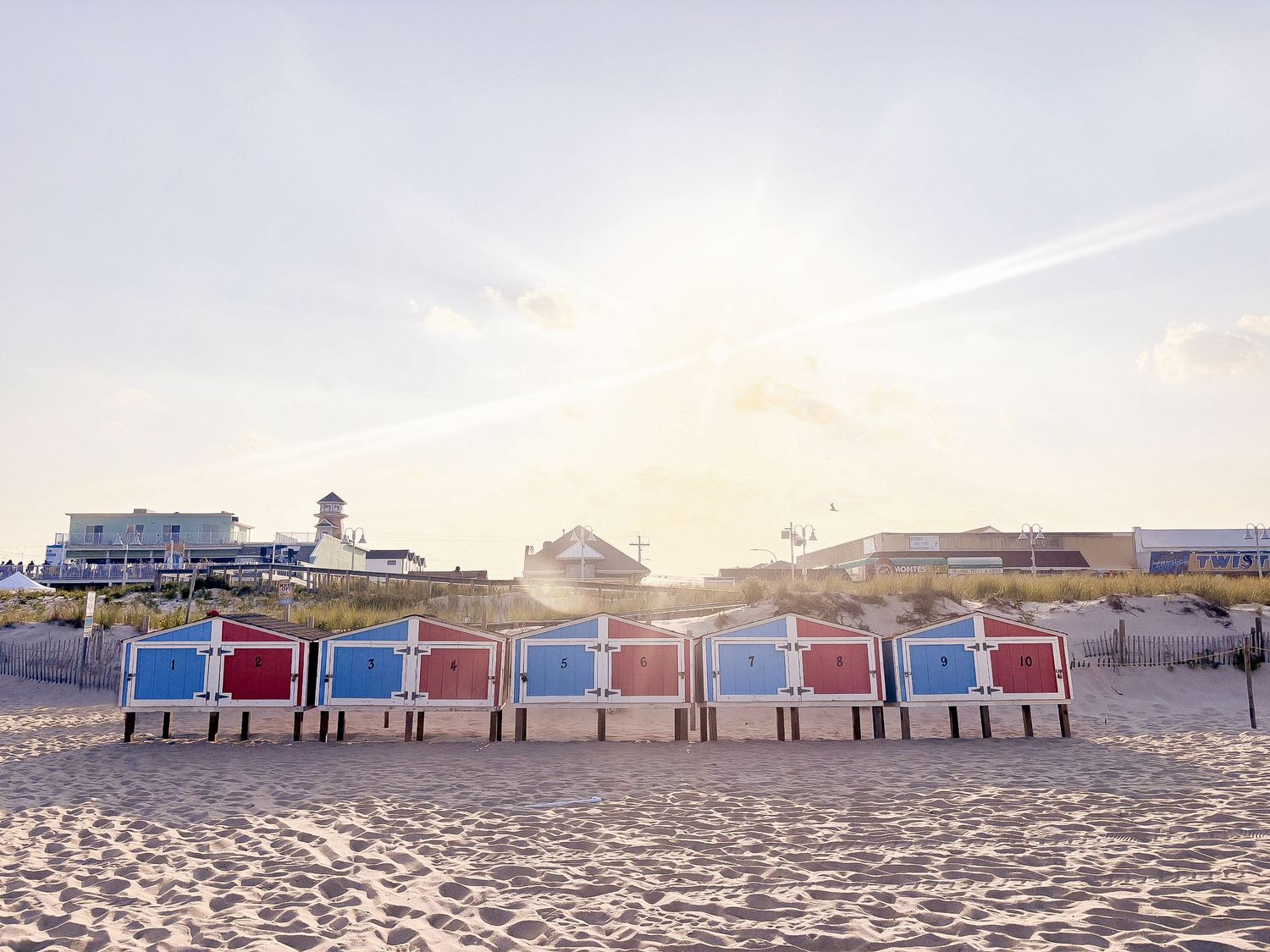 Colorful beach cabins lined up on sandy shore with lighthouse and buildings under bright sunlight.