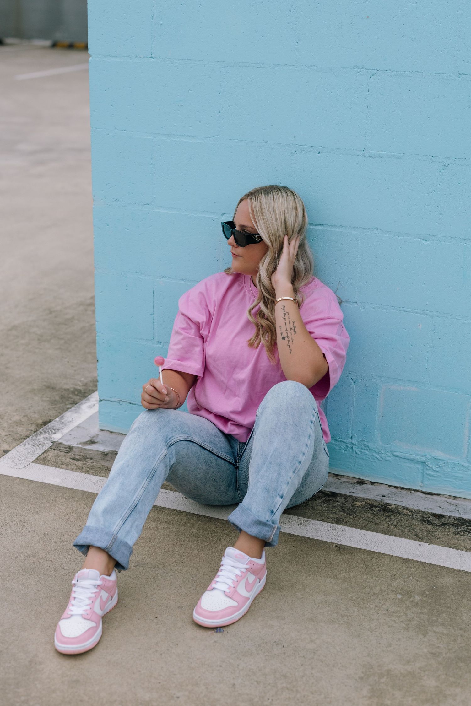 Casual street style photo of someone in pink shirt and jeans sitting against a blue wall.