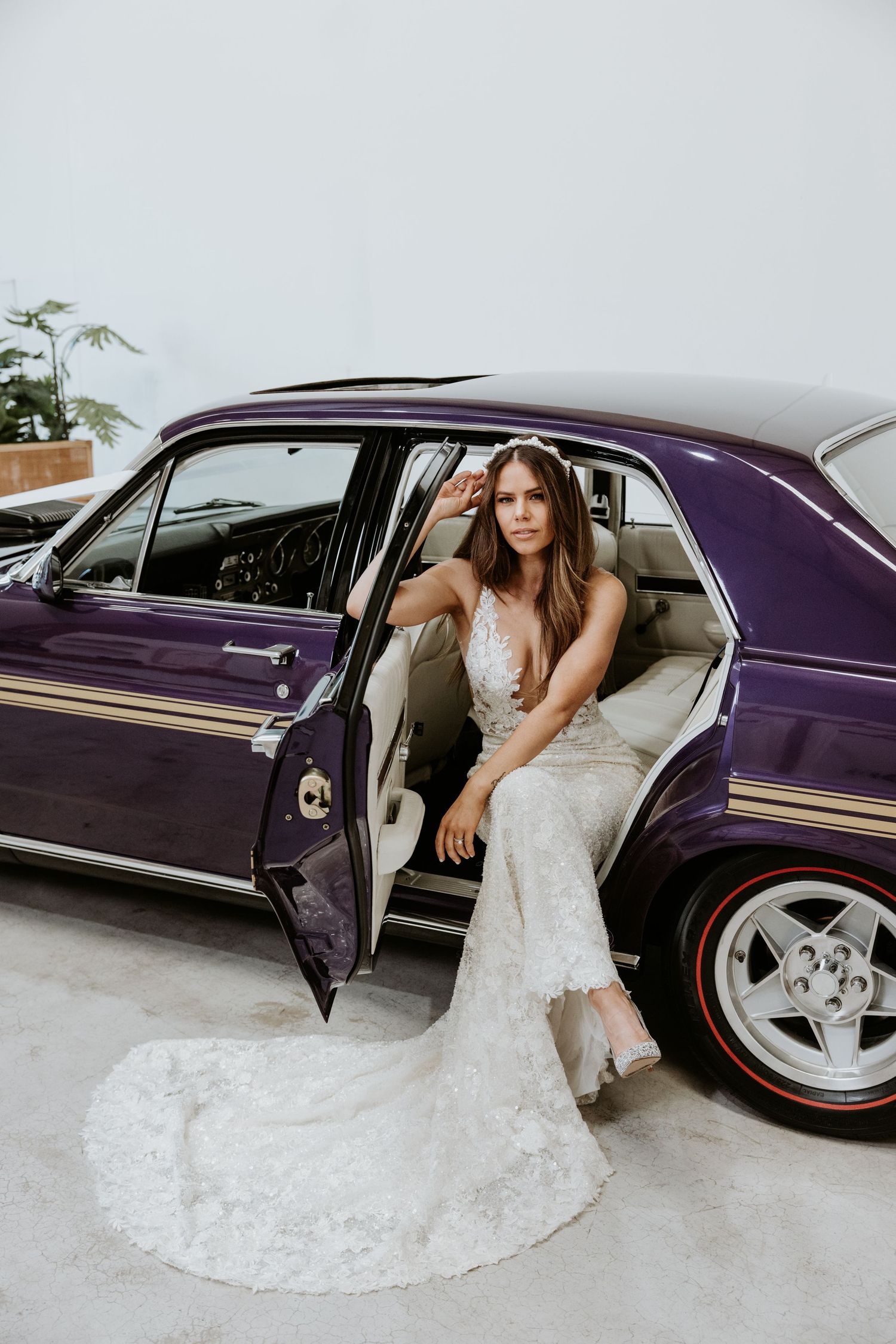 Bride in lace wedding gown sitting in open door of vintage purple classic car for elegant bridal photo shoot.