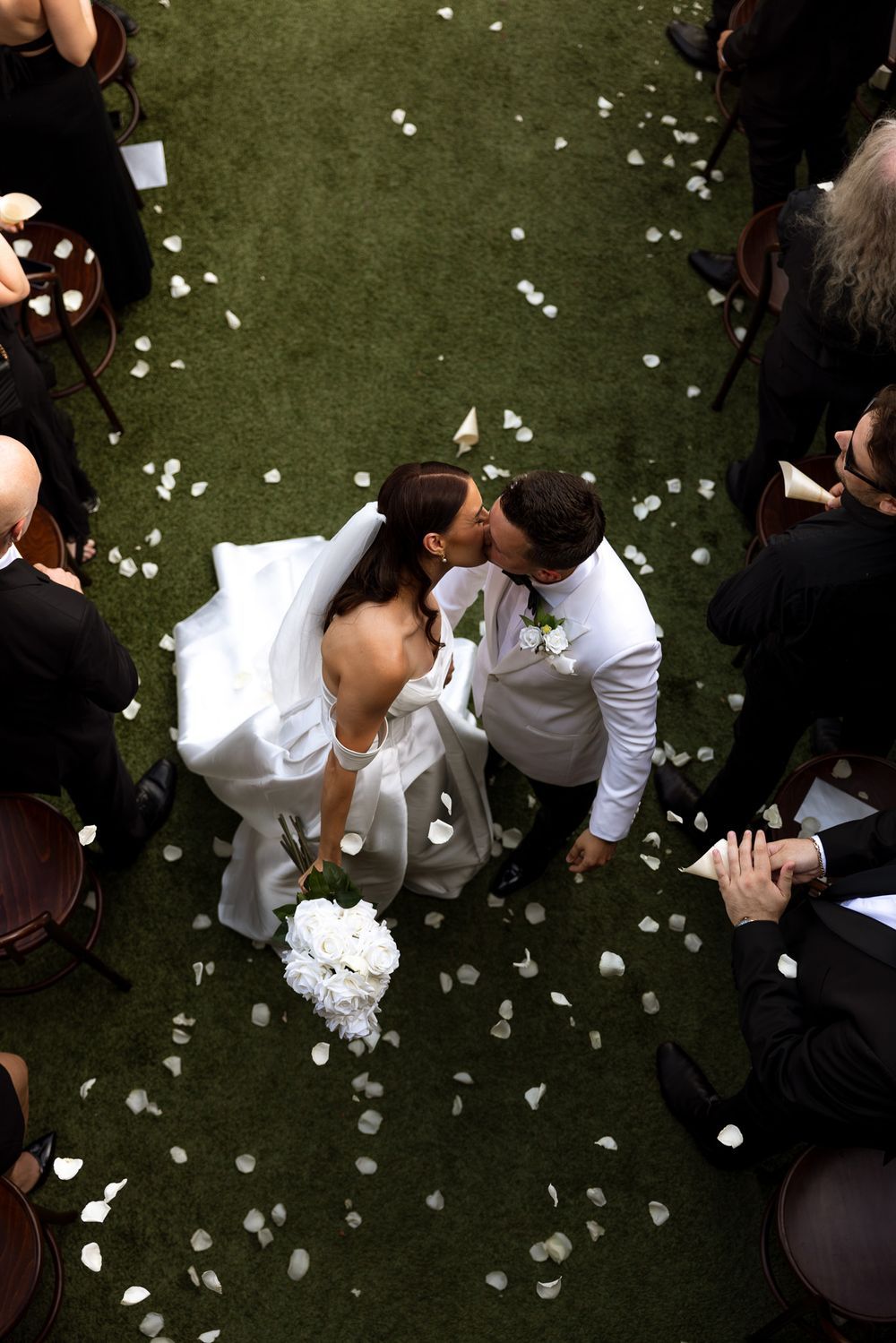 Aerial view of wedding couple lying on grass surrounded by scattered white flower petals.
