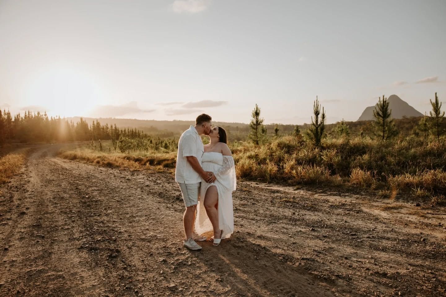 Couple sharing a romantic moment on a dirt road at golden sunset with lush greenery and mountains in the background.