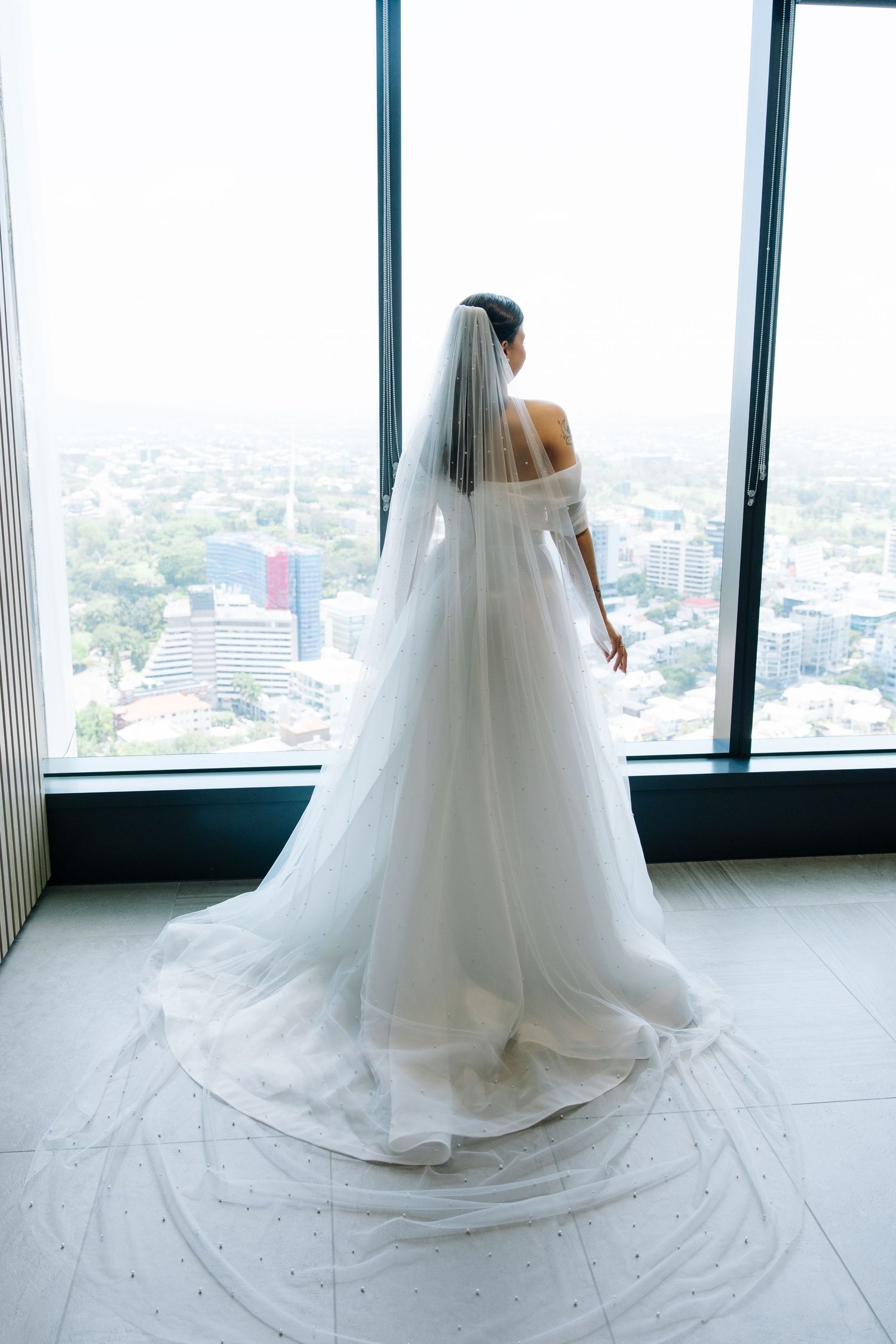 Bride in flowing white gown and veil looking out floor-to-ceiling windows at city view.