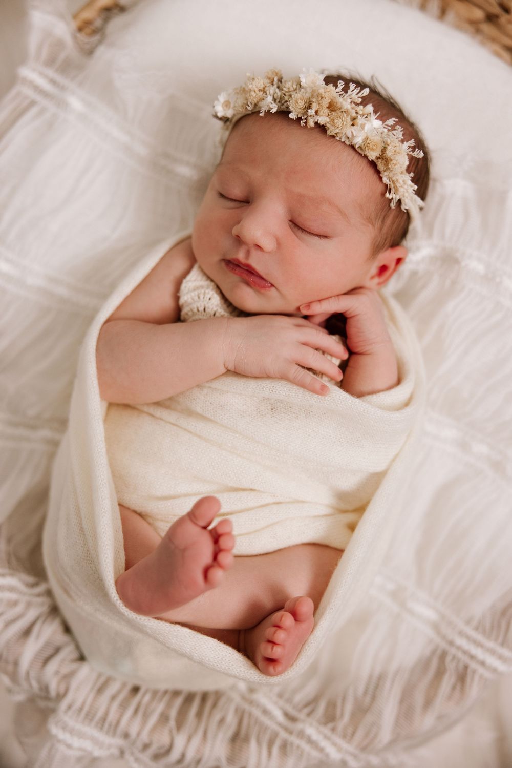 Peaceful sleeping newborn wearing delicate floral crown and white wrap on cream bedding.