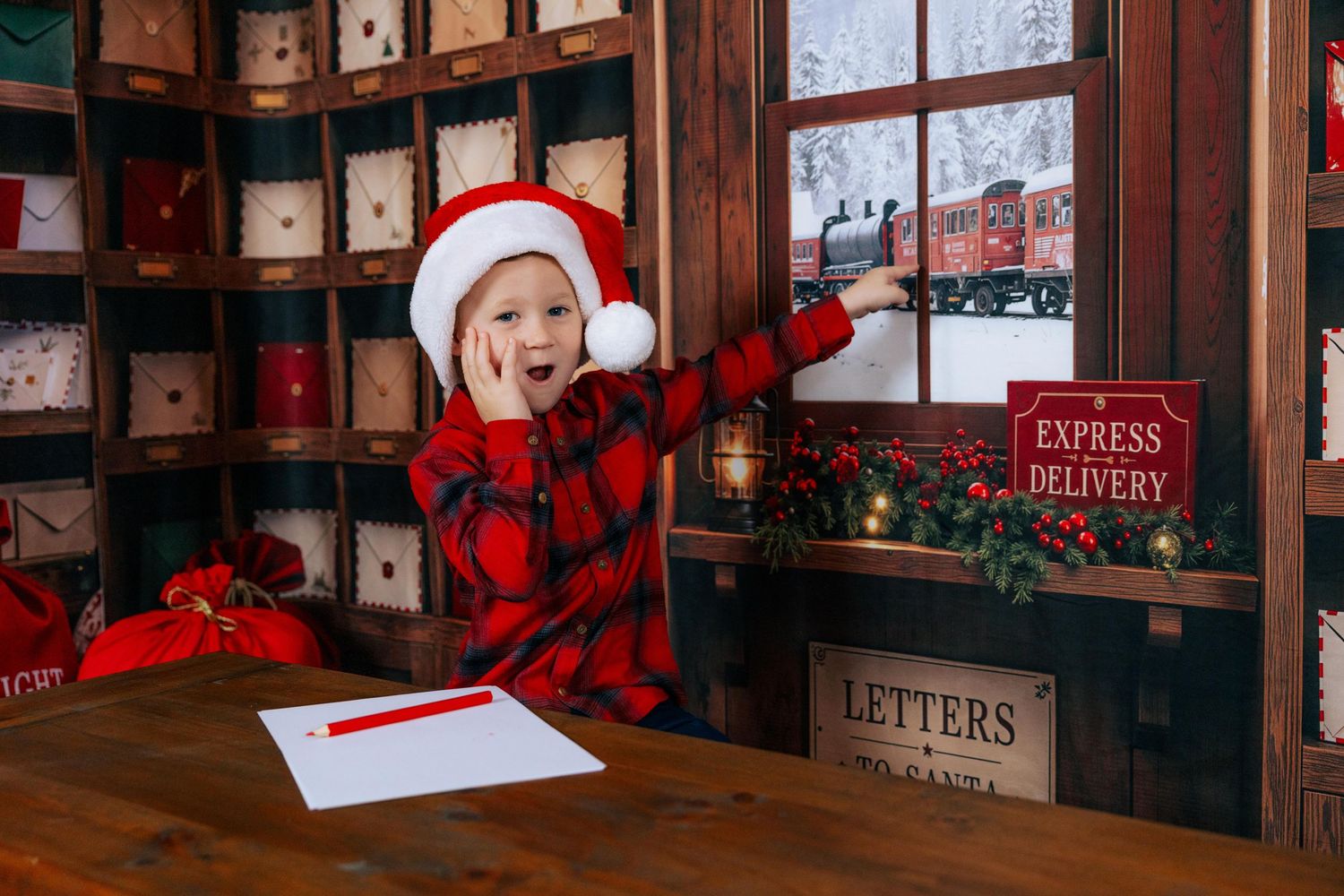 Child in Santa hat and plaid pajamas writing a letter to Santa in a festive Christmas post office setting.