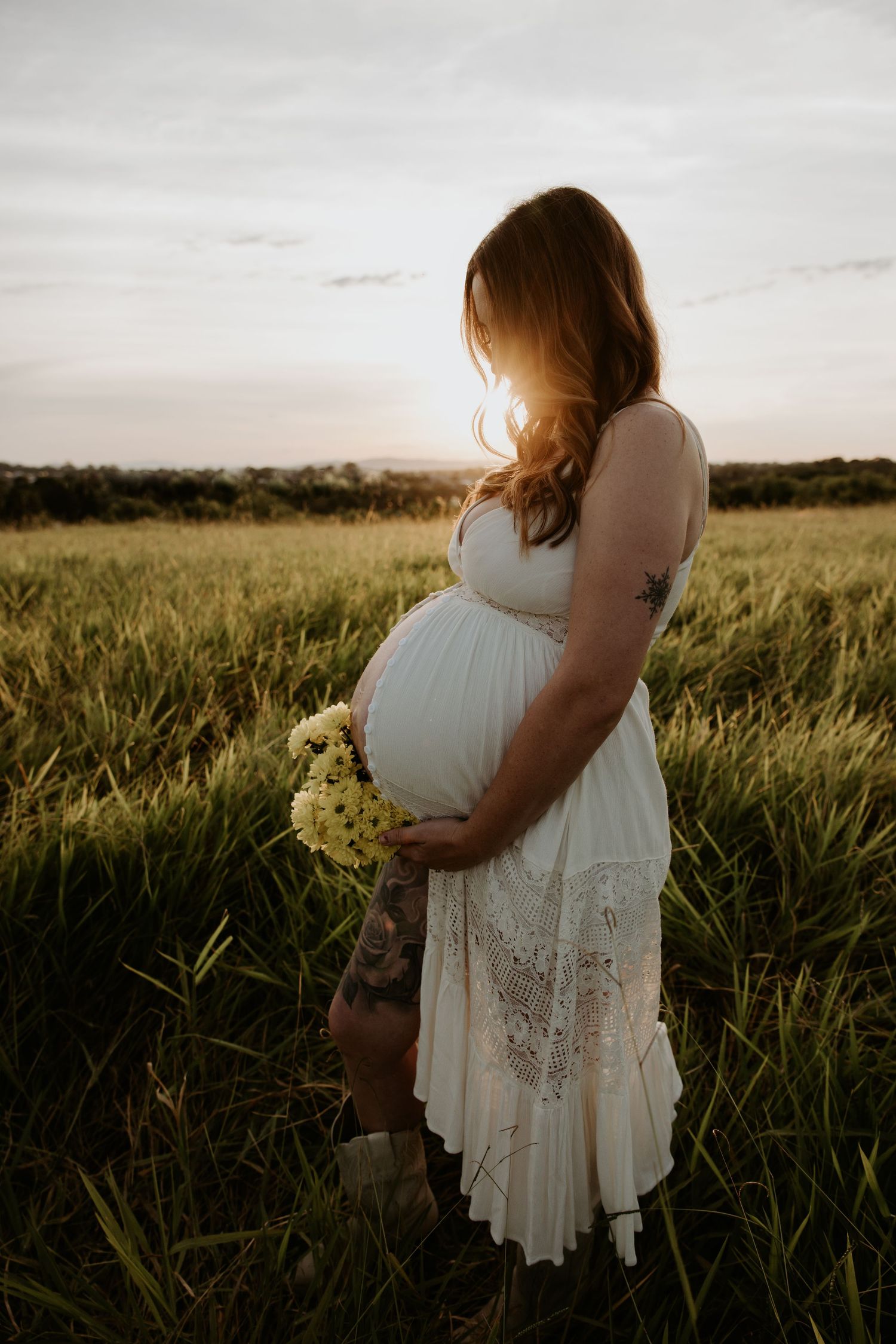 Silhouette of pregnant woman in white dress standing in sunlit field.