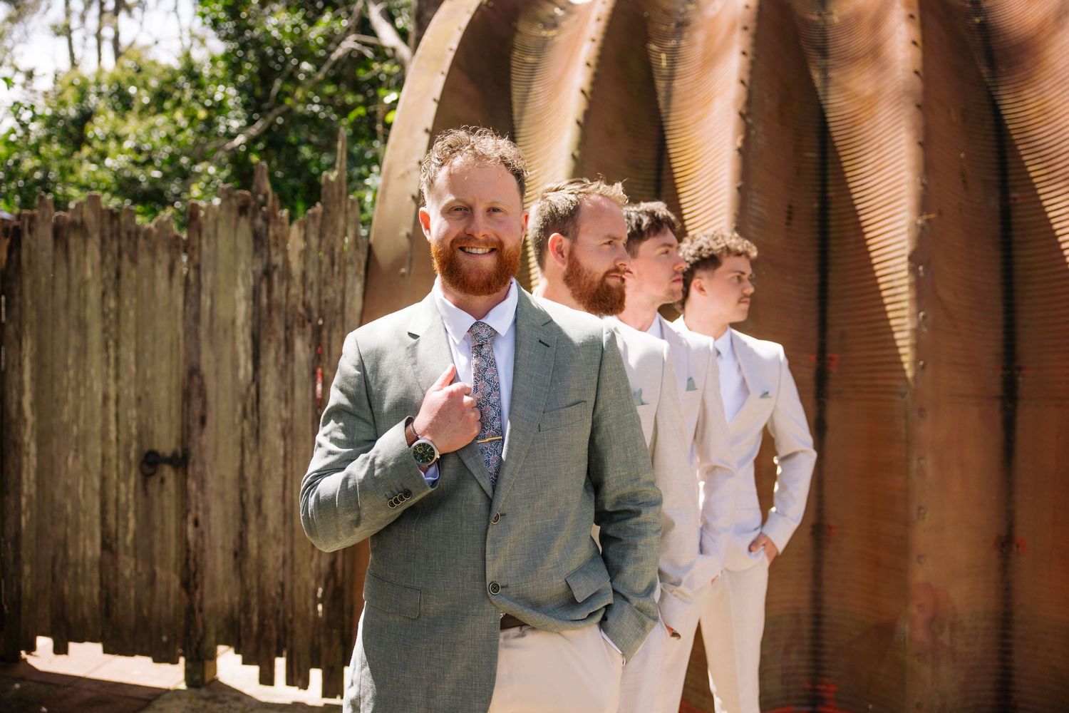 Groomsmen in light colored suits standing against rustic wall outdoors.