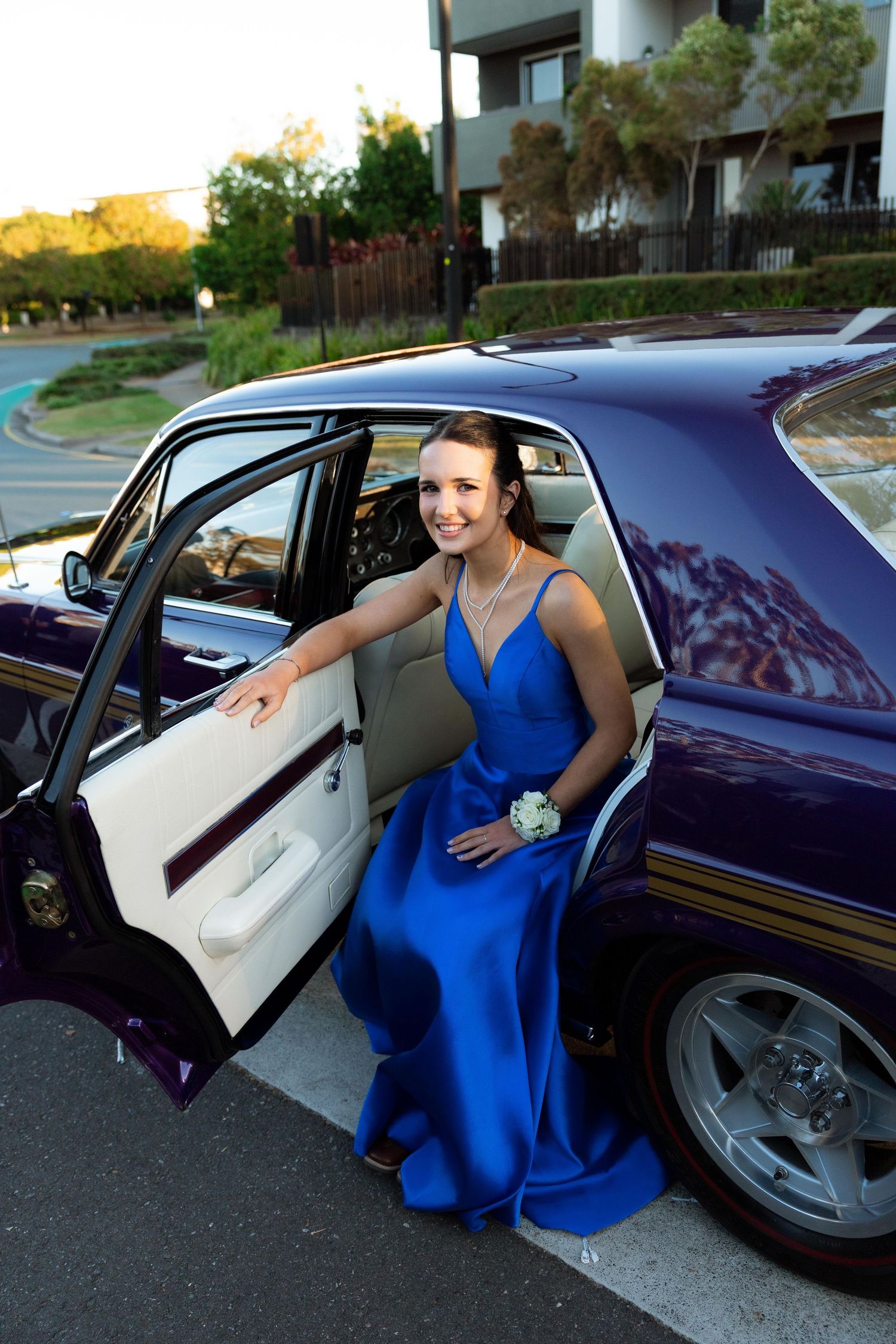 Young woman in royal blue prom dress stepping out of a purple classic car, wearing a wrist corsage.