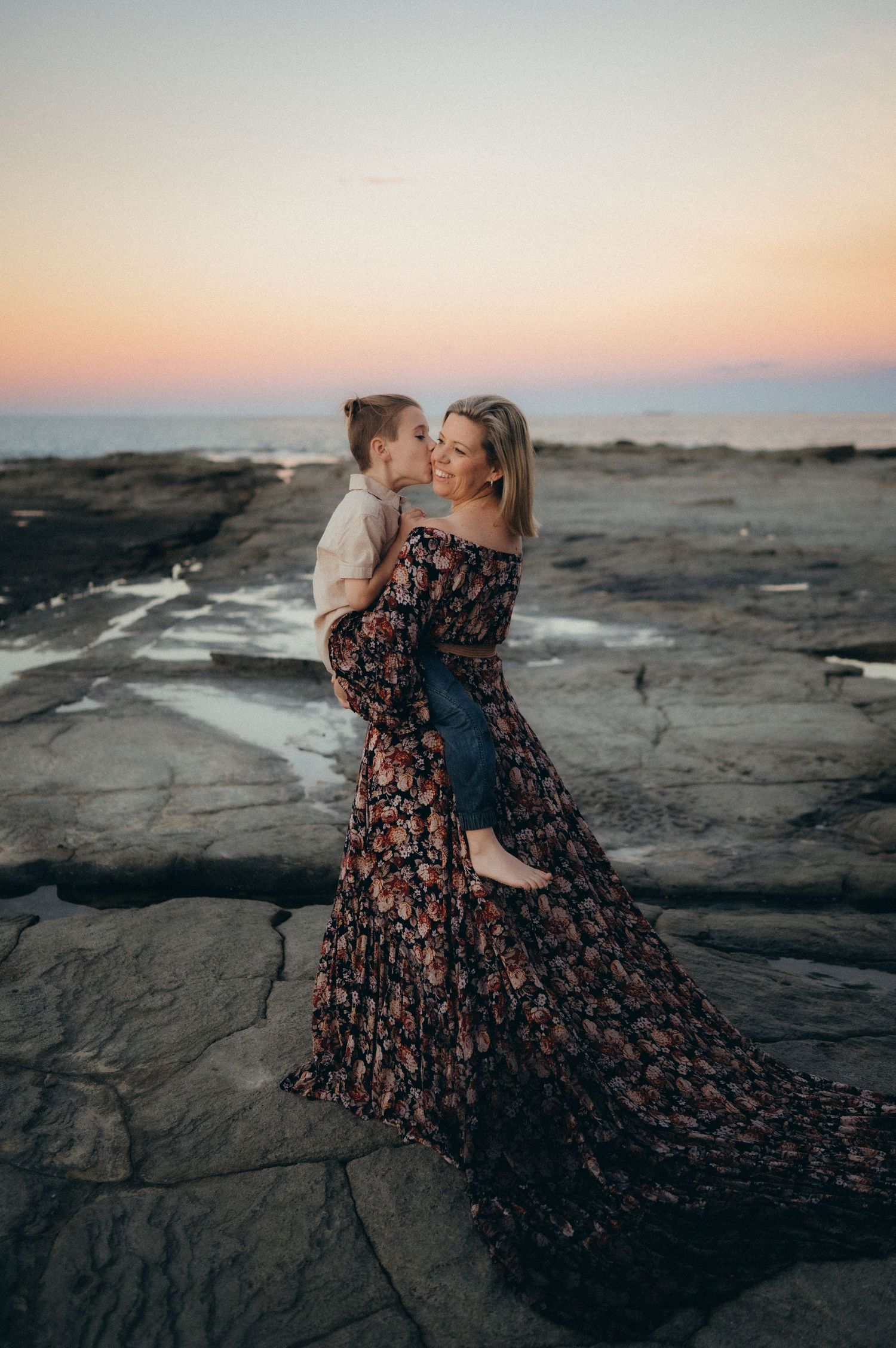 A romantic sunset scene on rocky coastal cliffs with a flowing floral dress catching the evening light.