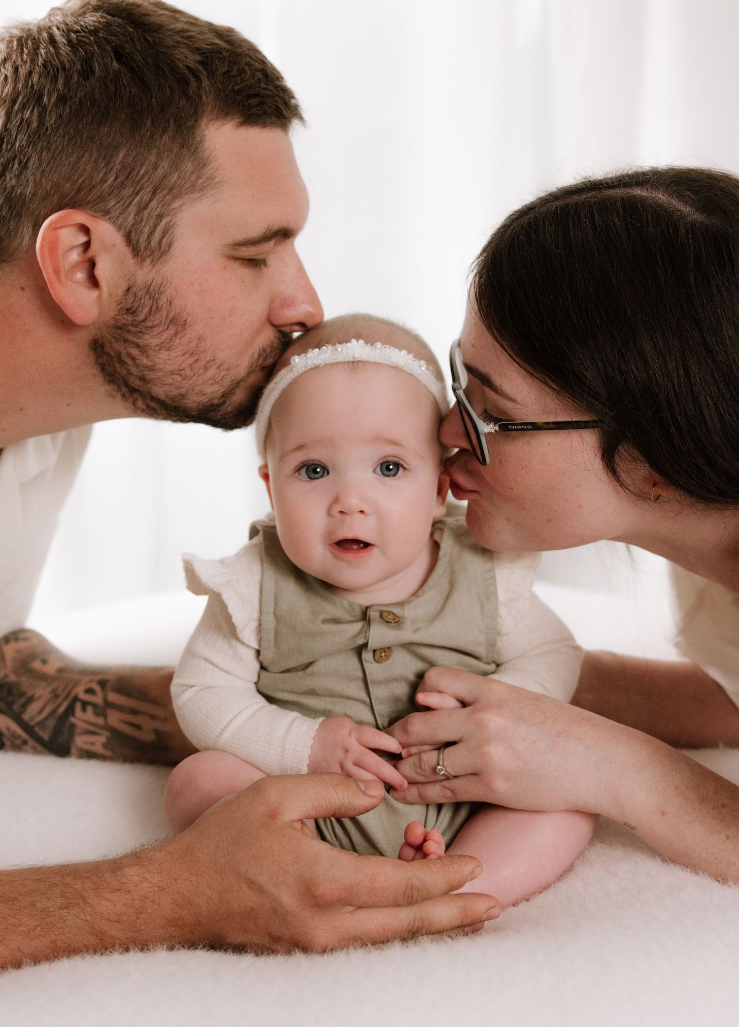 Sweet family moment with baby getting kisses from parents on white background.