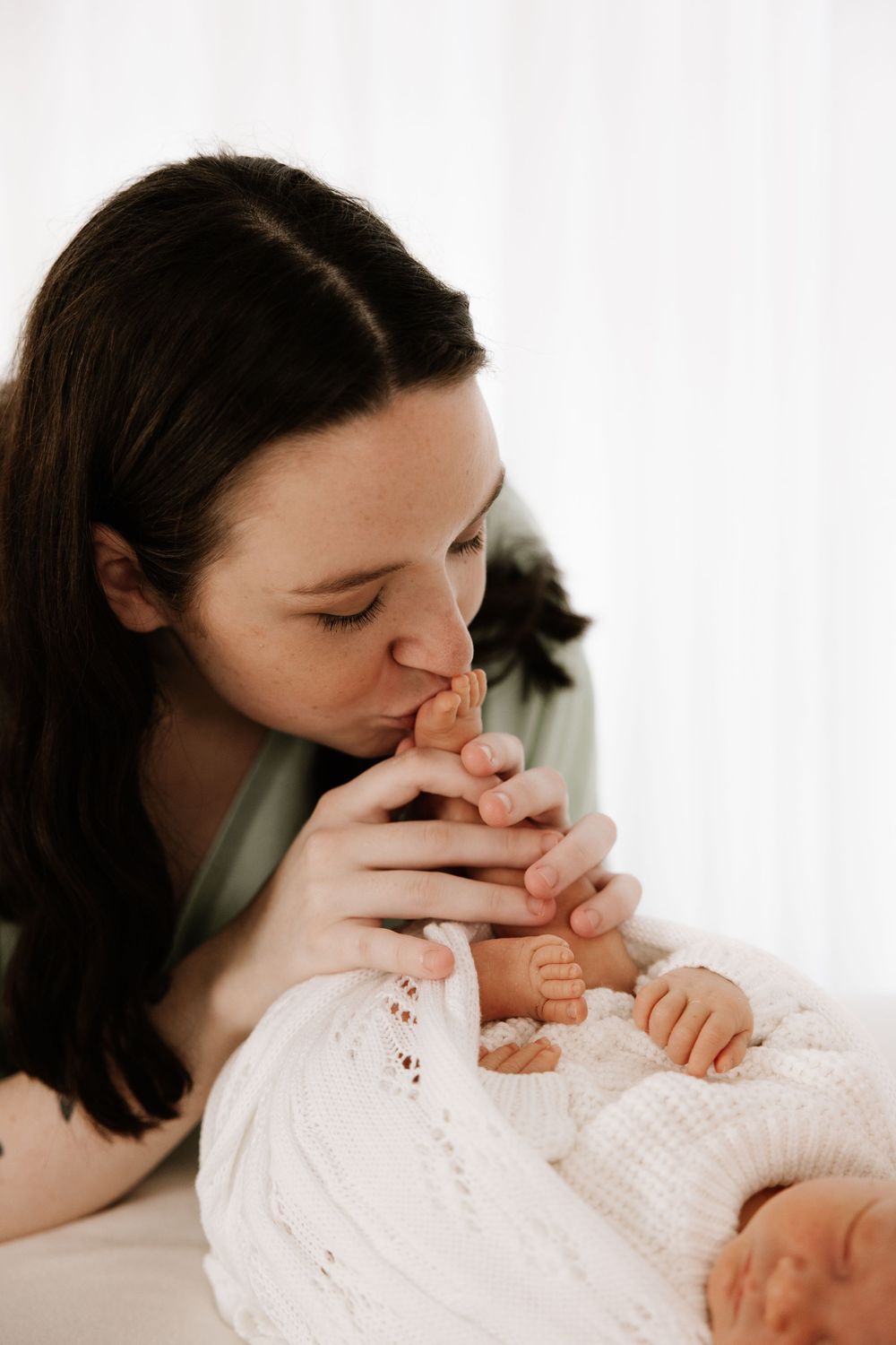 Tender closeup of gentle kisses and cuddles with sleeping newborn in white.