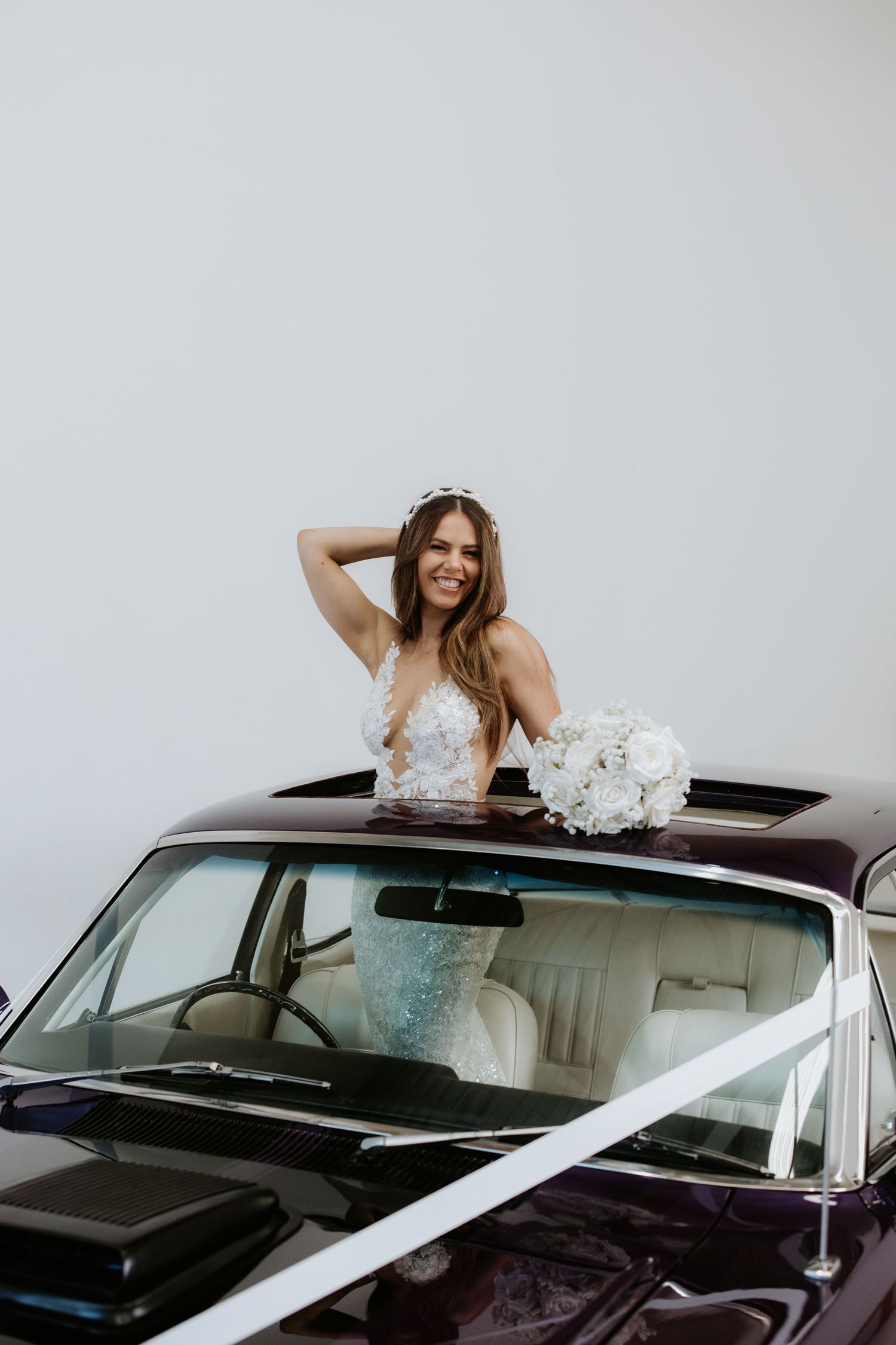 Smiling bride in white gown holding bouquet, posing through sunroof of decorated vintage dark car.