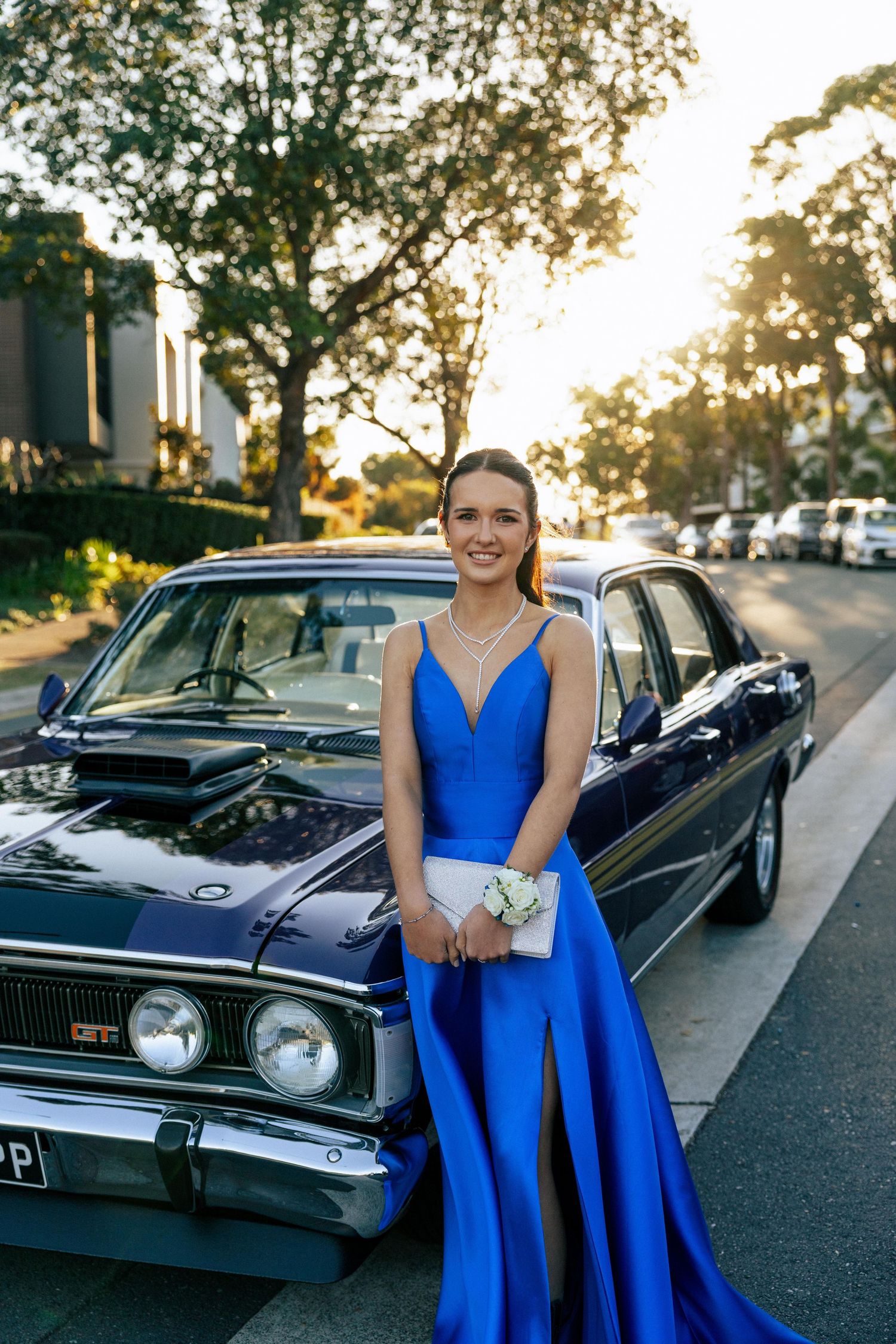 Young woman in royal blue prom gown poses beside a classic dark blue sedan on a tree-lined street at golden hour.