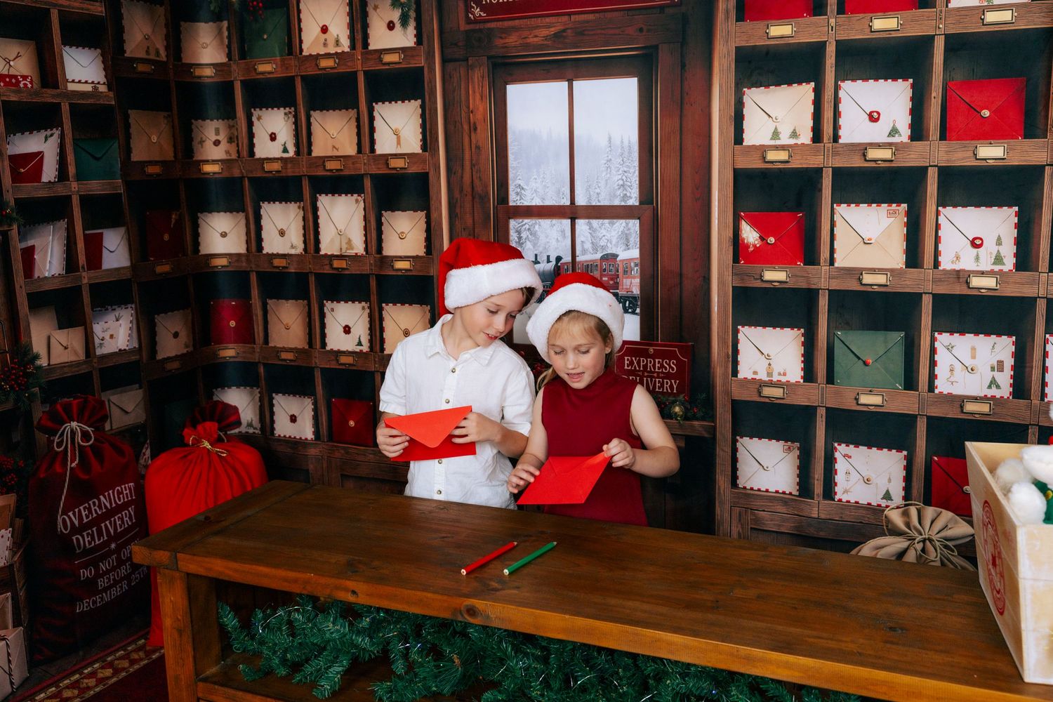Two children in Santa hats write Christmas letters at a wooden desk surrounded by festive mailboxes and holiday decorations.
