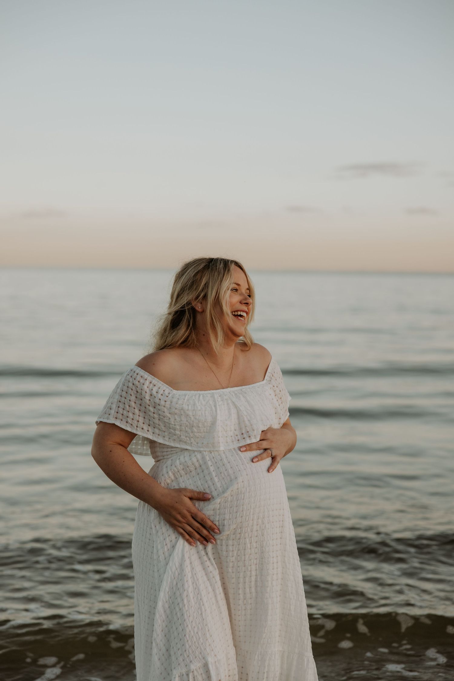 Woman in flowing white dress laughs joyfully by the ocean at dusk.