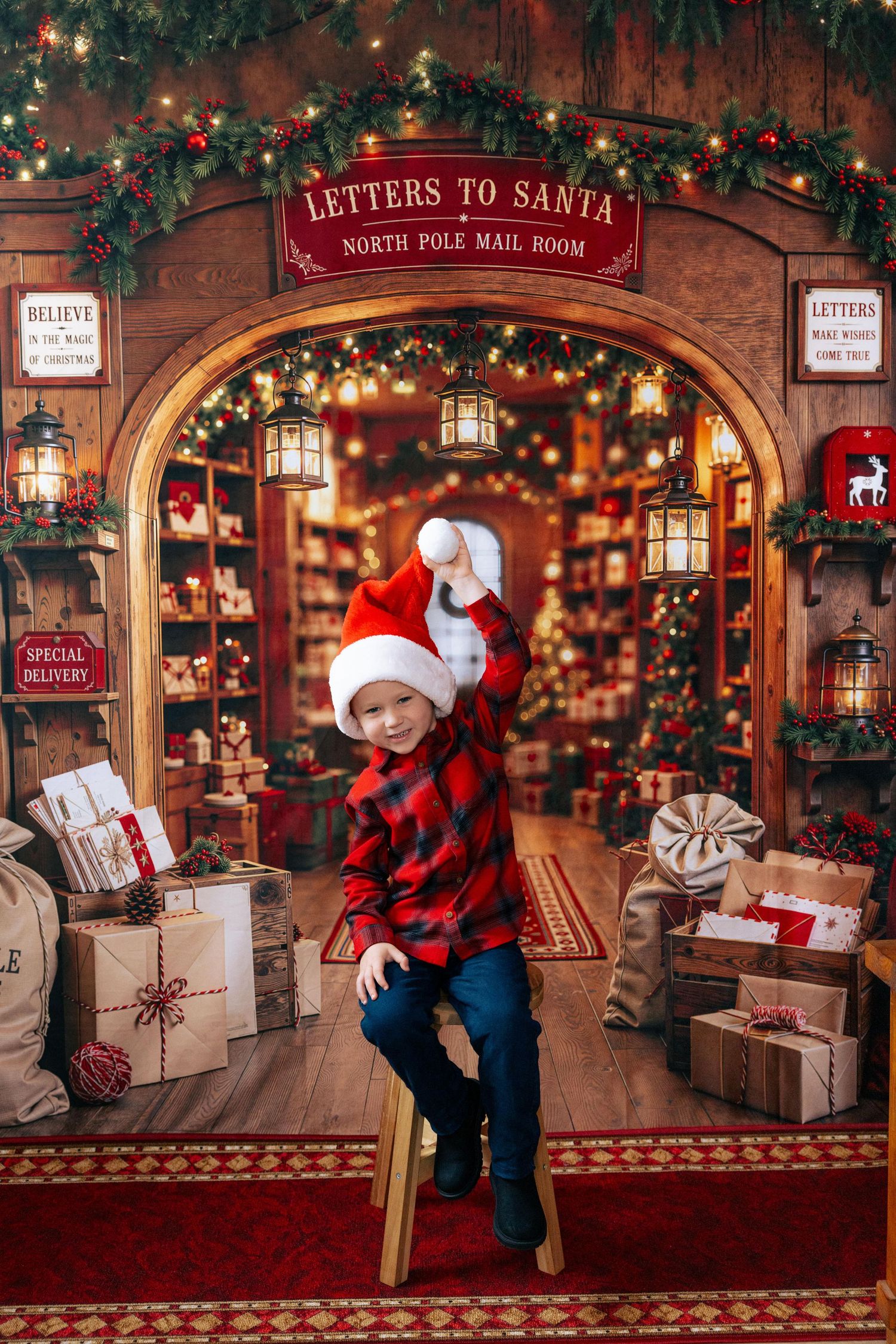 A cheerful boy in a Santa hat and plaid shirt sits before a festive North Pole mail room Christmas backdrop.