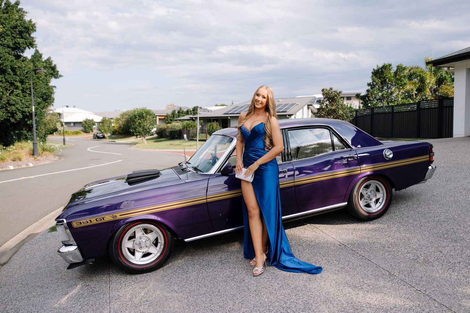 Woman in blue gown posing beside a classic purple muscle car in a parking lot.
