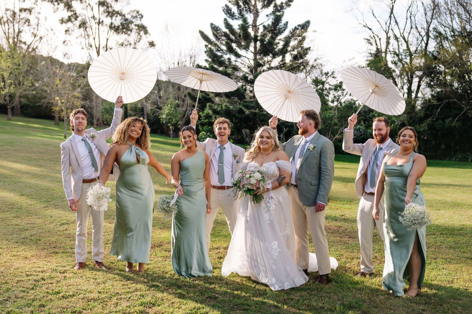 Wedding party in mint green and light colored attire holding white parasols on a sunny lawn.