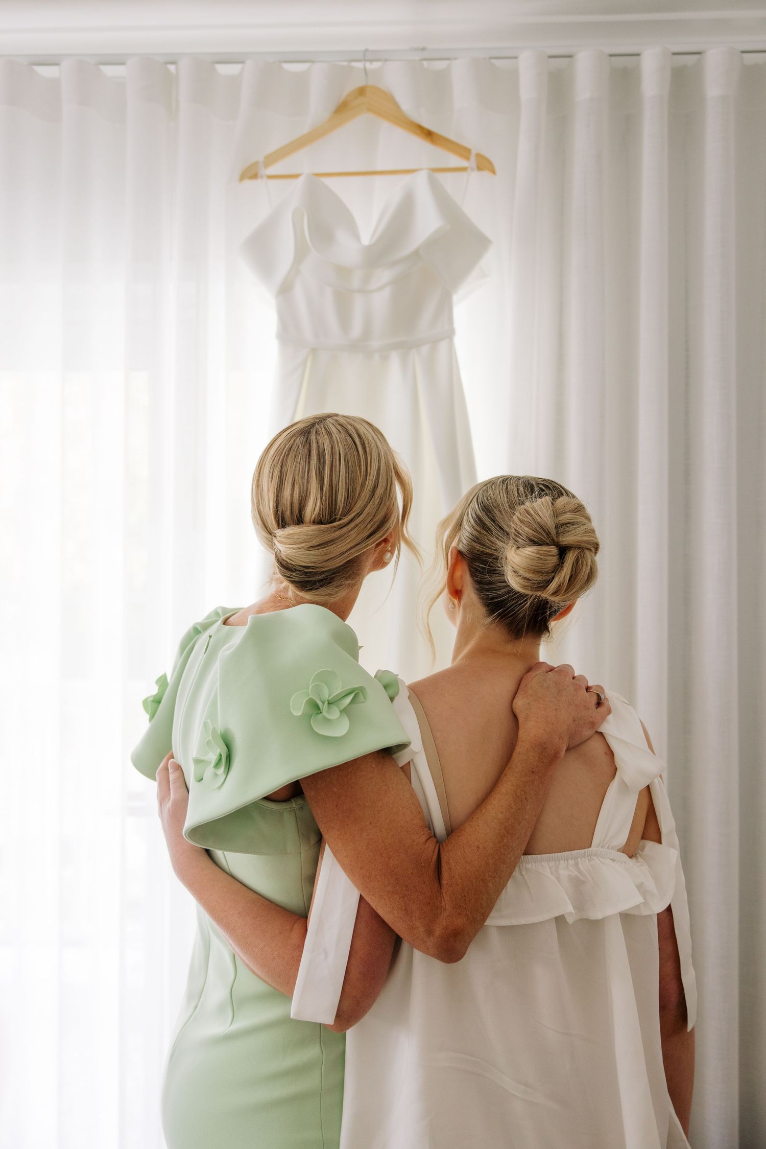 Bridesmaids in mint green dresses help with wedding preparations against white curtains.