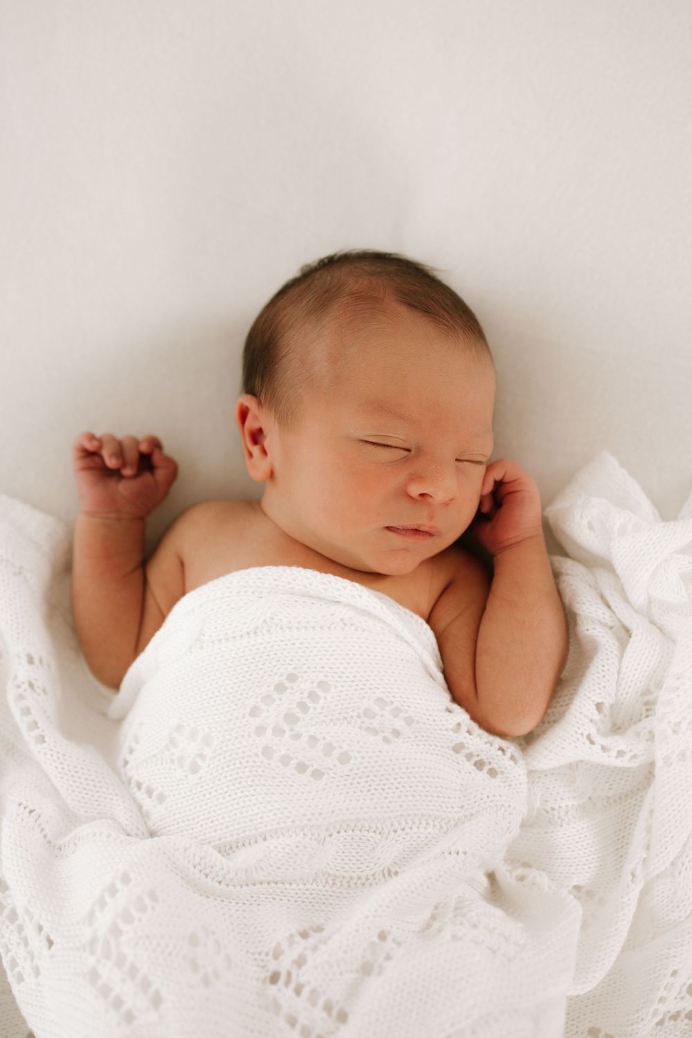 Sleeping newborn baby wrapped in delicate white lace blanket against light background.