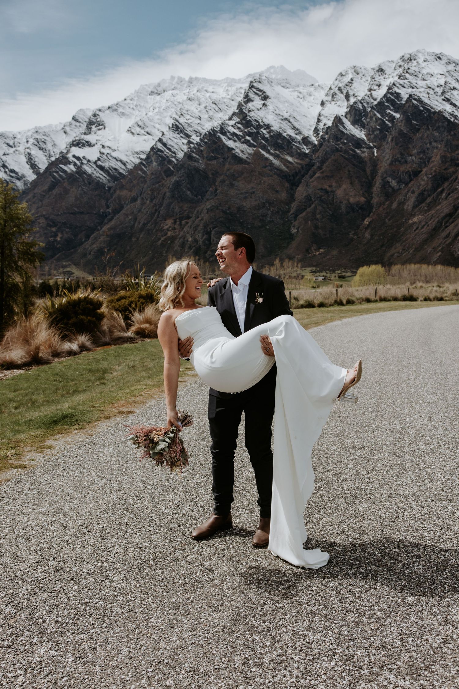 Wedding couple performs dramatic dip poses on gravel path with mountain landscape behind them.