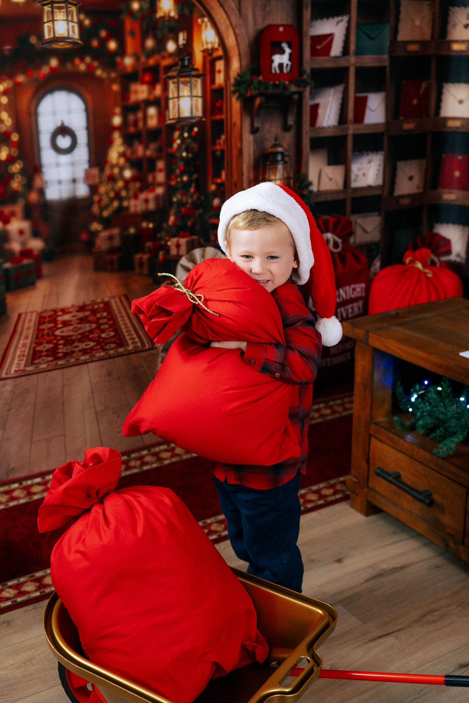 Toddler in Santa hat hugging large red gift sack in festive Christmas workshop with decorated shelves.