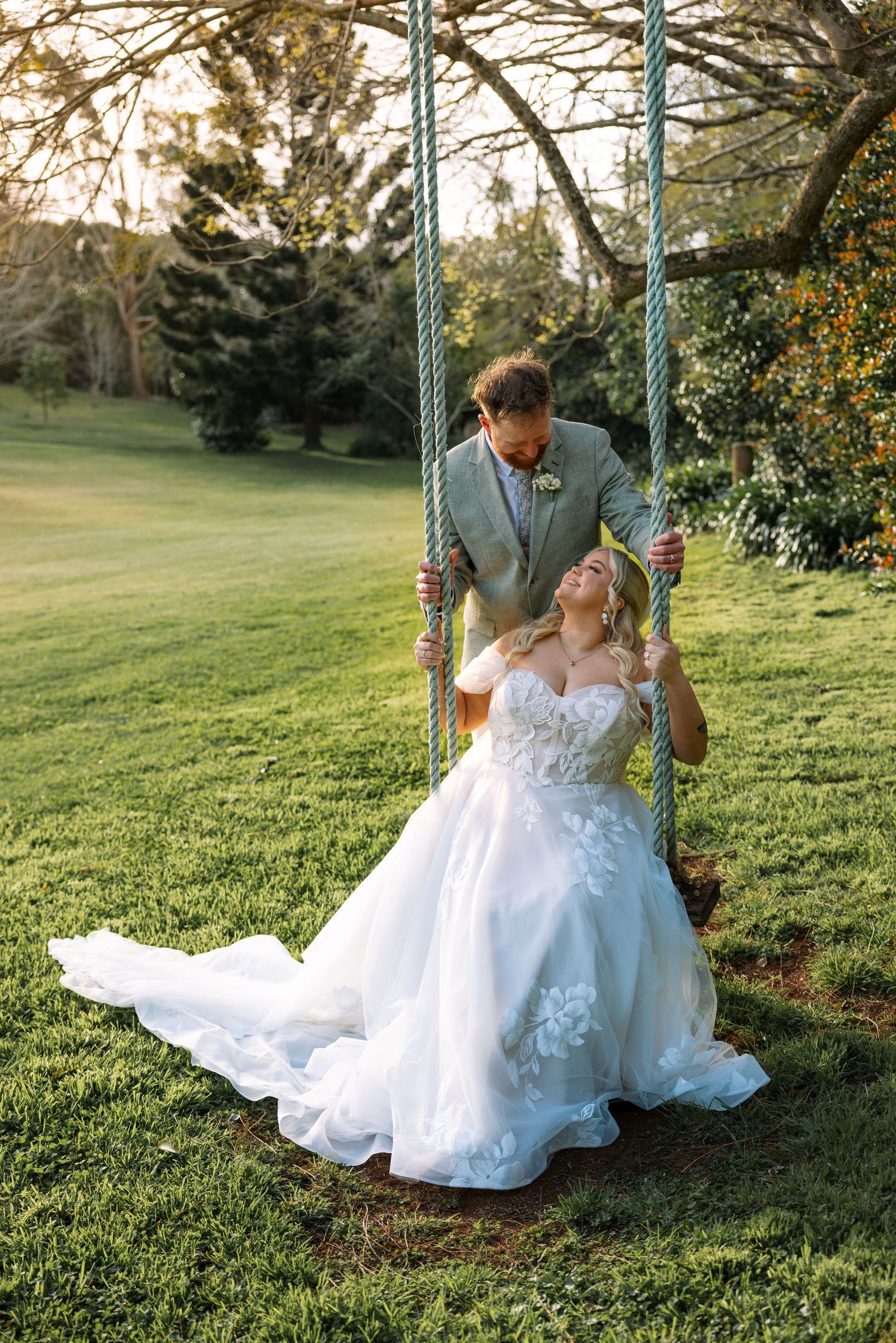 Bride on decorative garden swing in flowing white lace dress against green landscape.