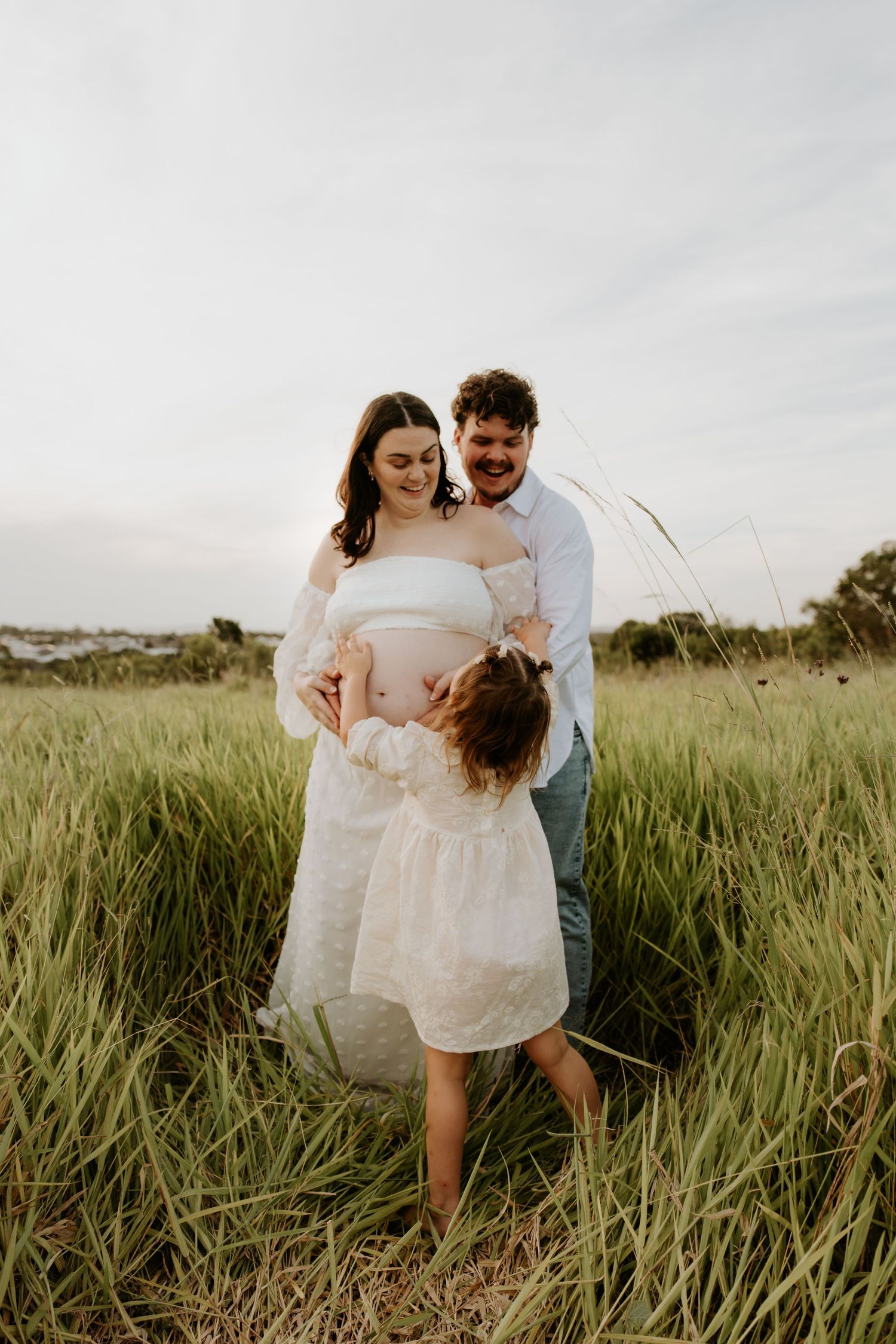 Pregnant woman in white dress with partner and young daughter in a lush green field during a maternity photoshoot.