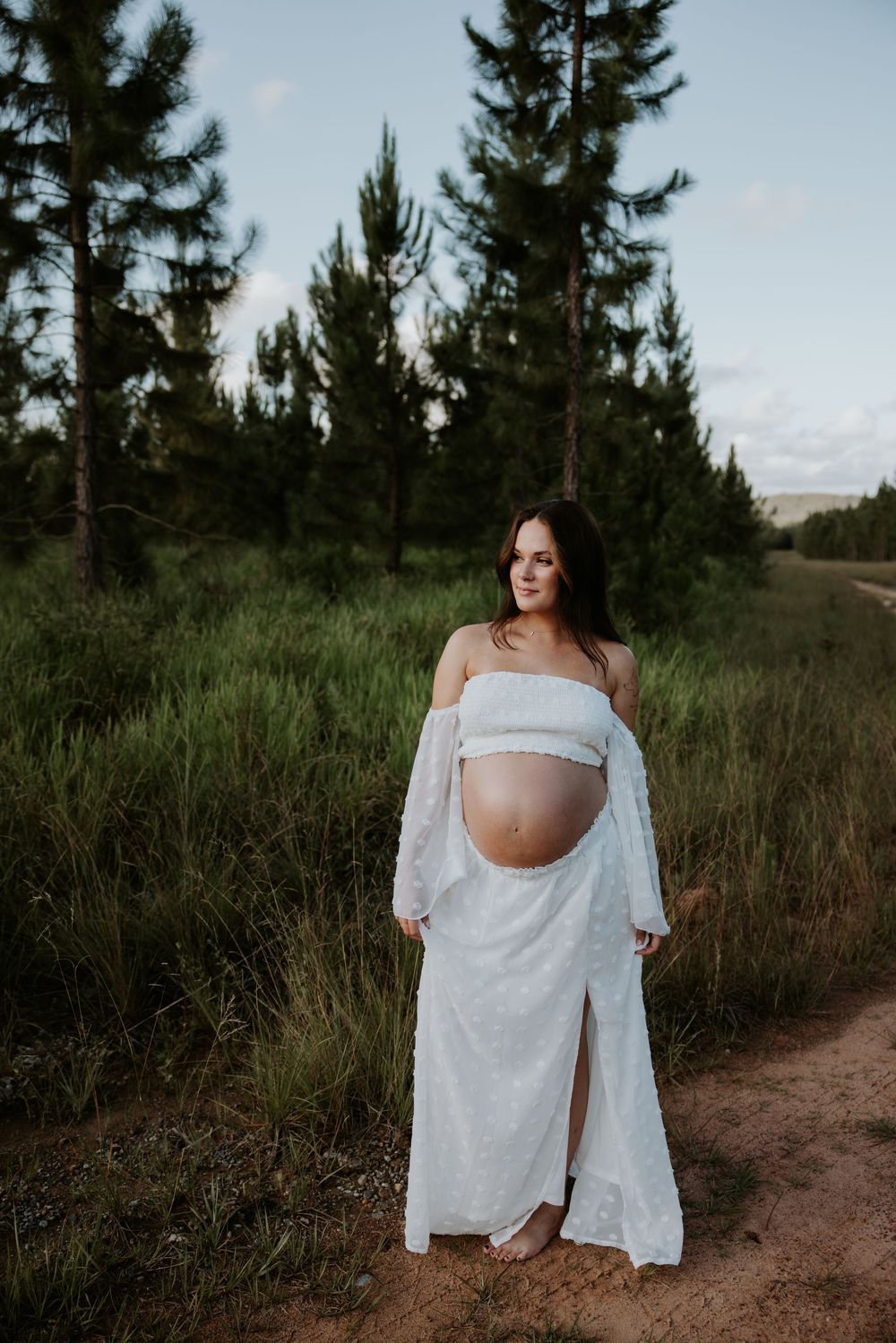 Maternity photoshoot in a rustic outdoor setting with pine trees and a dirt path at sunset.