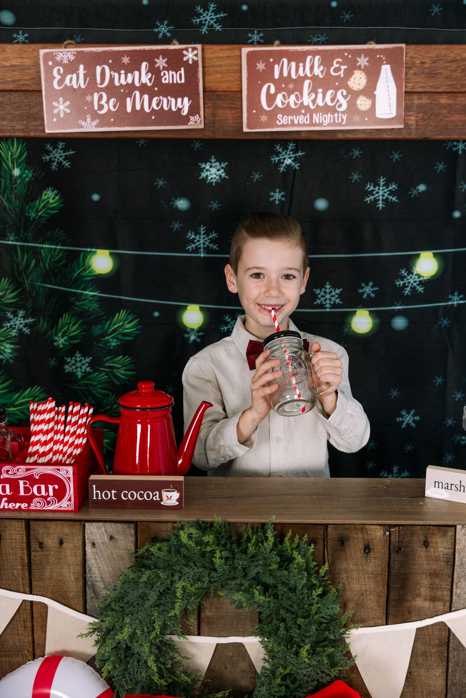 Christmas photo booth with hot cocoa bar, string lights and rustic wooden signs displaying festive messages.
