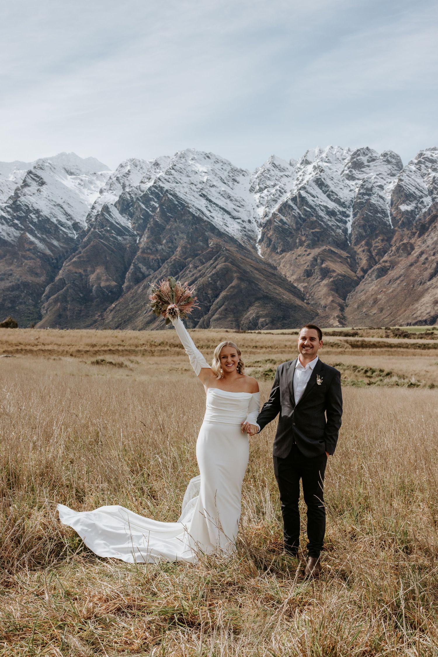Wedding couple poses dramatically in a field with snow-capped mountains creating a stunning backdrop.