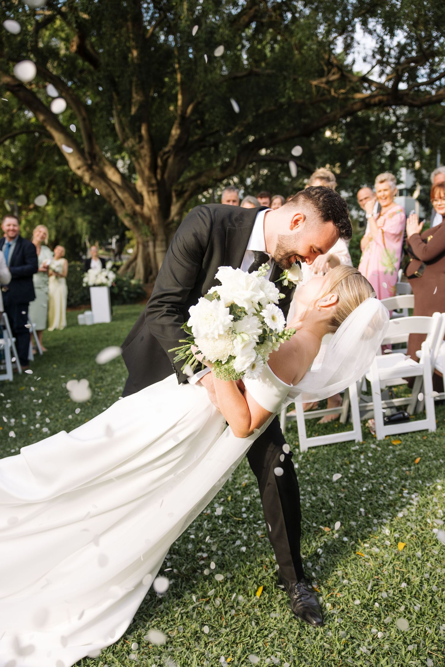 Playful wedding dip pose on grass lawn with guests watching in background.