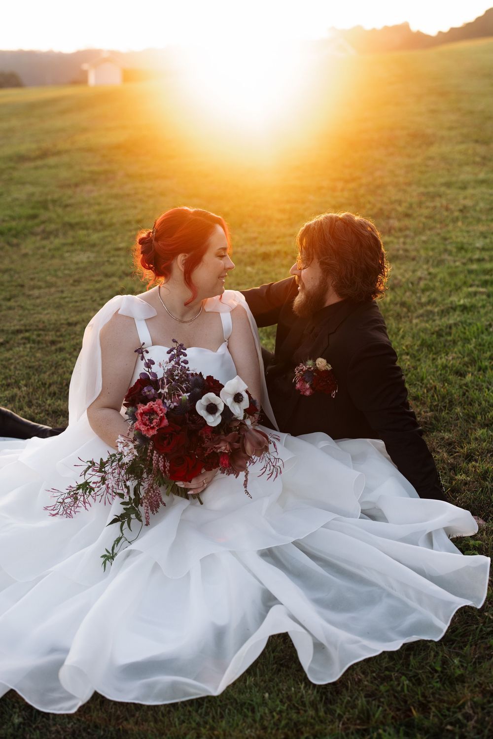 Two people in white dresses share intimate moment at sunset with burgundy flowers.
