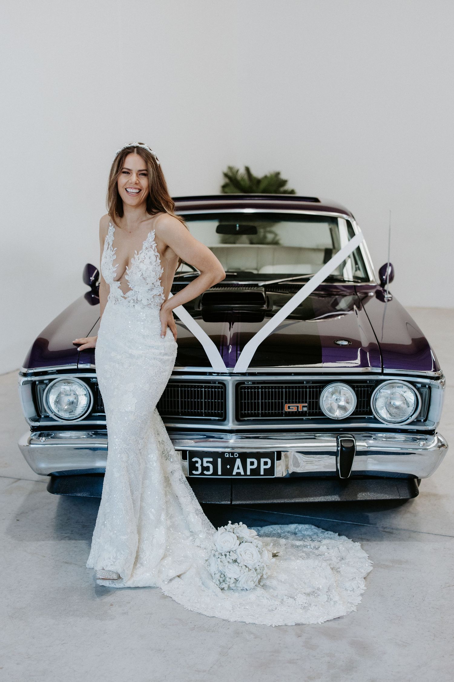 Bride in lace mermaid wedding gown poses in front of a purple vintage GTC car decorated with white ribbons.