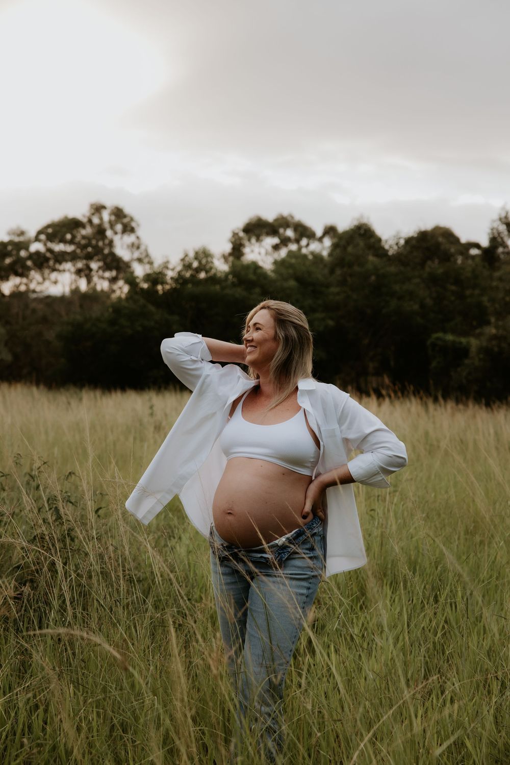 Expectant mother in white shirt and jeans posing confidently in grassy field.