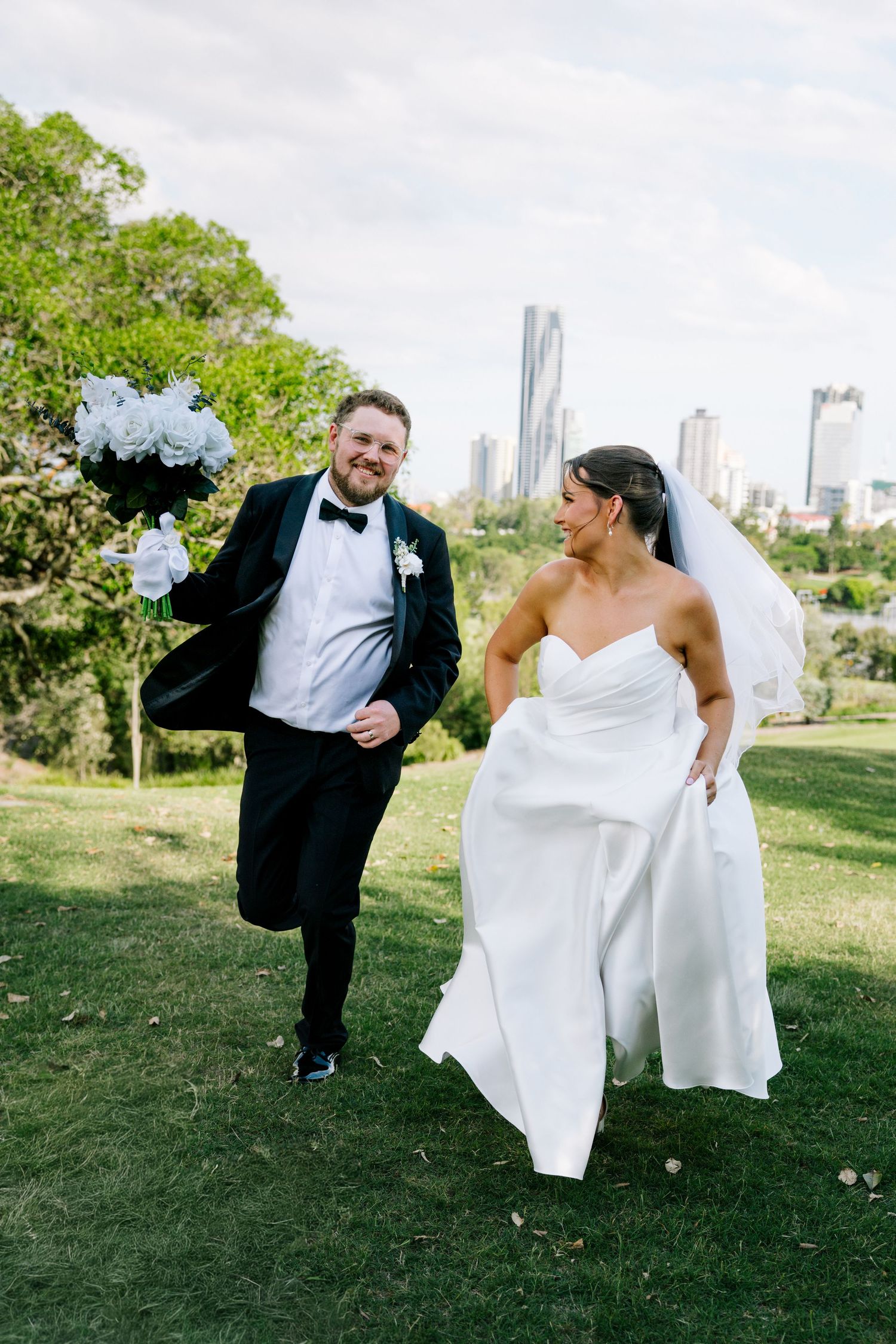 A playful wedding couple dances and poses on a grassy lawn with city buildings in the background.