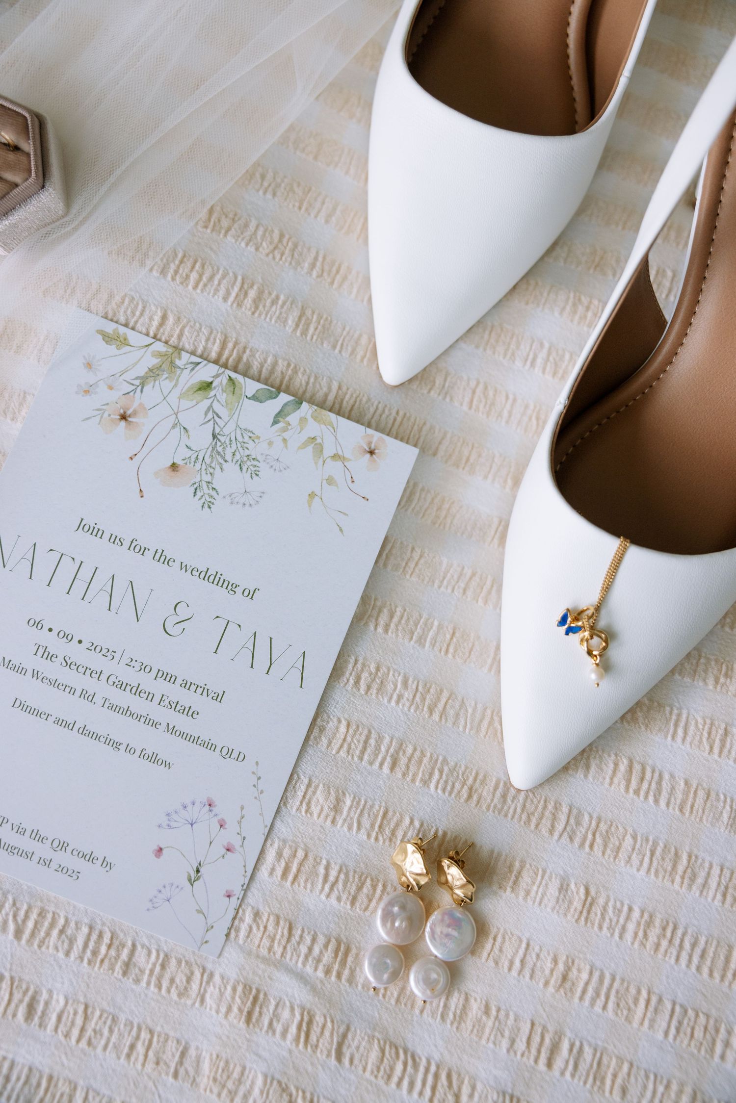 White pointed bridal shoes with floral wedding invitation and pearl earrings on textured fabric.