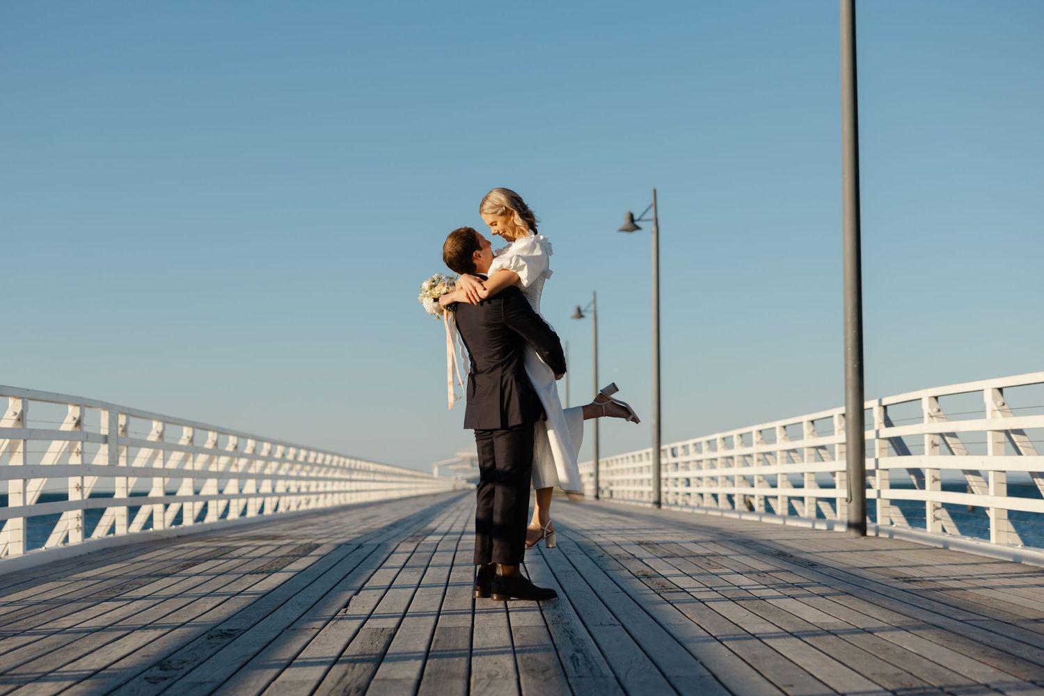 Groom lifting bride on a sunny pier with white railings and clear blue sky on their wedding day.