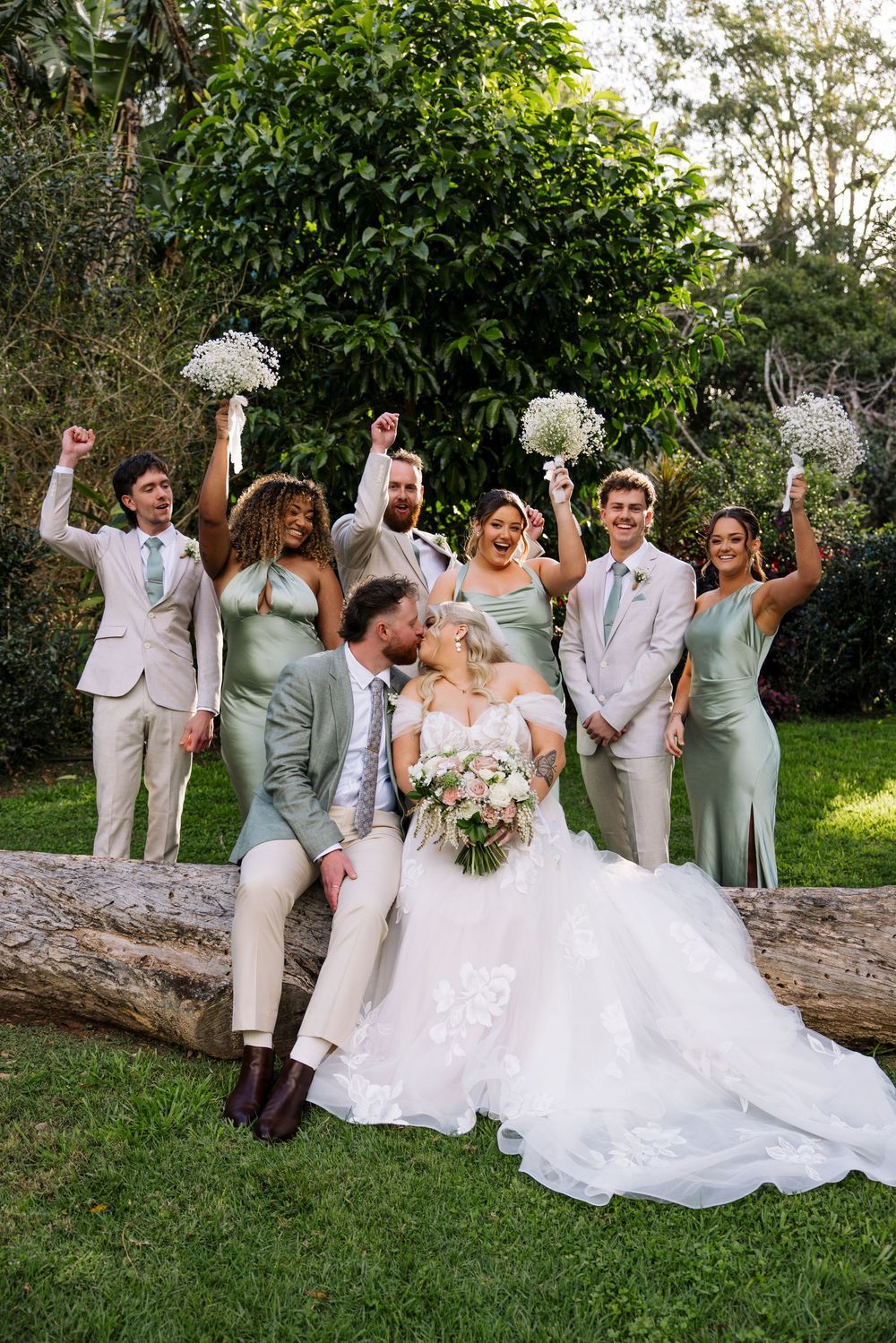 Wedding party in mint green dresses and light gray suits poses casually on fallen log outdoors.