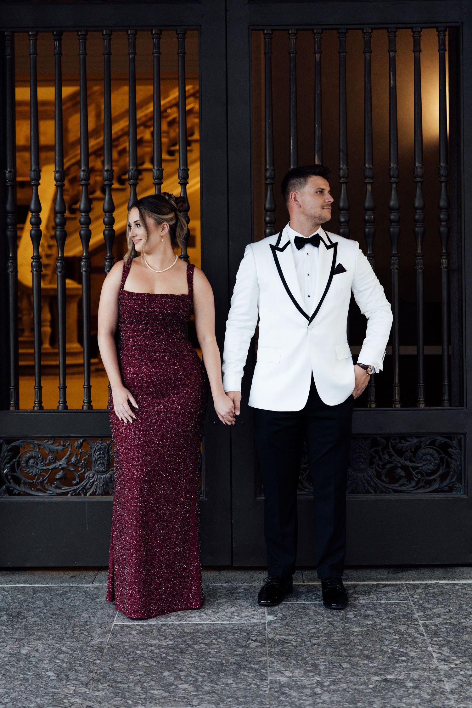Couple in formal attire posing before ornate black iron gates, woman in burgundy gown, man in white tuxedo jacket.