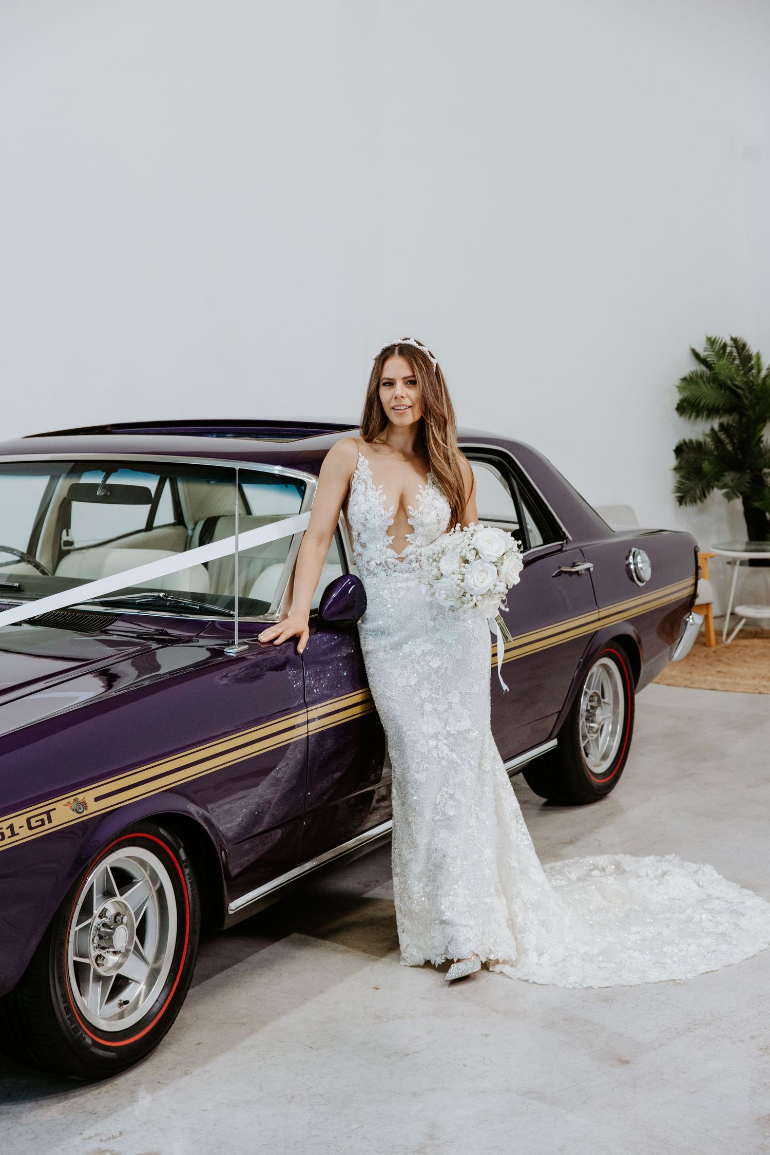 Bride in white lace gown holding bouquet poses beside a classic purple muscle car with gold stripes.