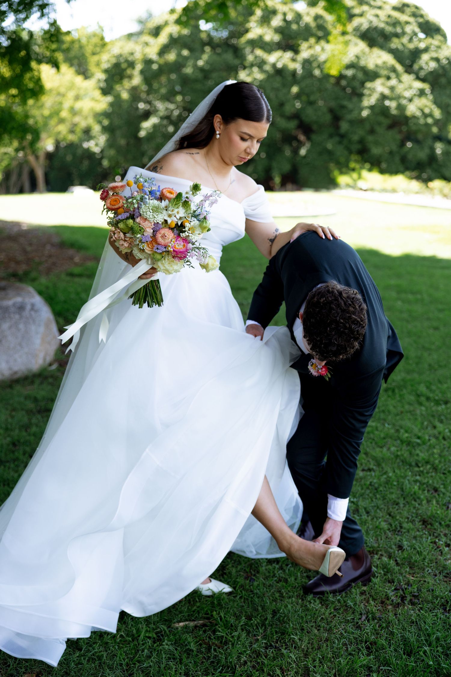 Groom helping bride with her shoe while she holds colorful bouquet outdoors.