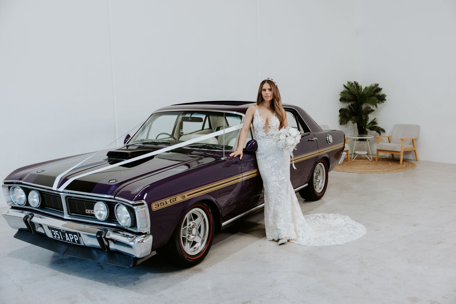 Bride in lace wedding gown posing beside a classic purple muscle car in a bright studio setting.